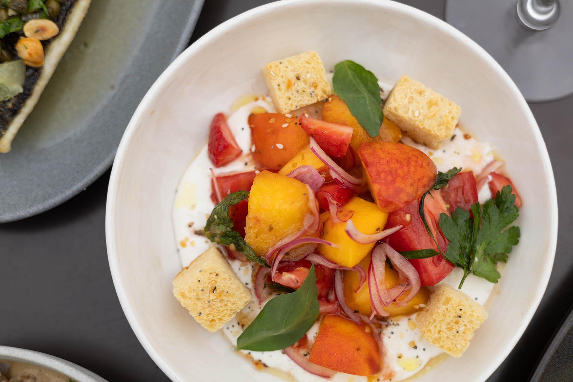 A white bowl filled with chunks of peach, tomato, croutons, red onions, fresh herbs, and a creamy base, garnished with basil and parsley.
