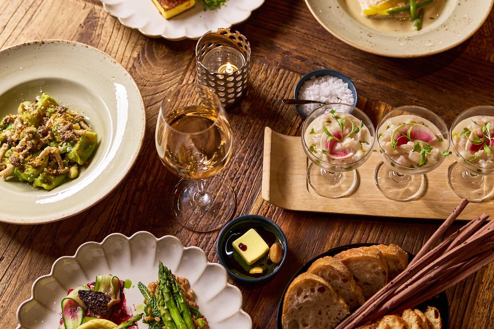 A wooden table set with assorted gourmet dishes, including pasta, vegetable plates, ceviche in glasses, a glass of white wine, bread slices, breadsticks, butter, and a bowl of salt, all lit by warm candlelight.