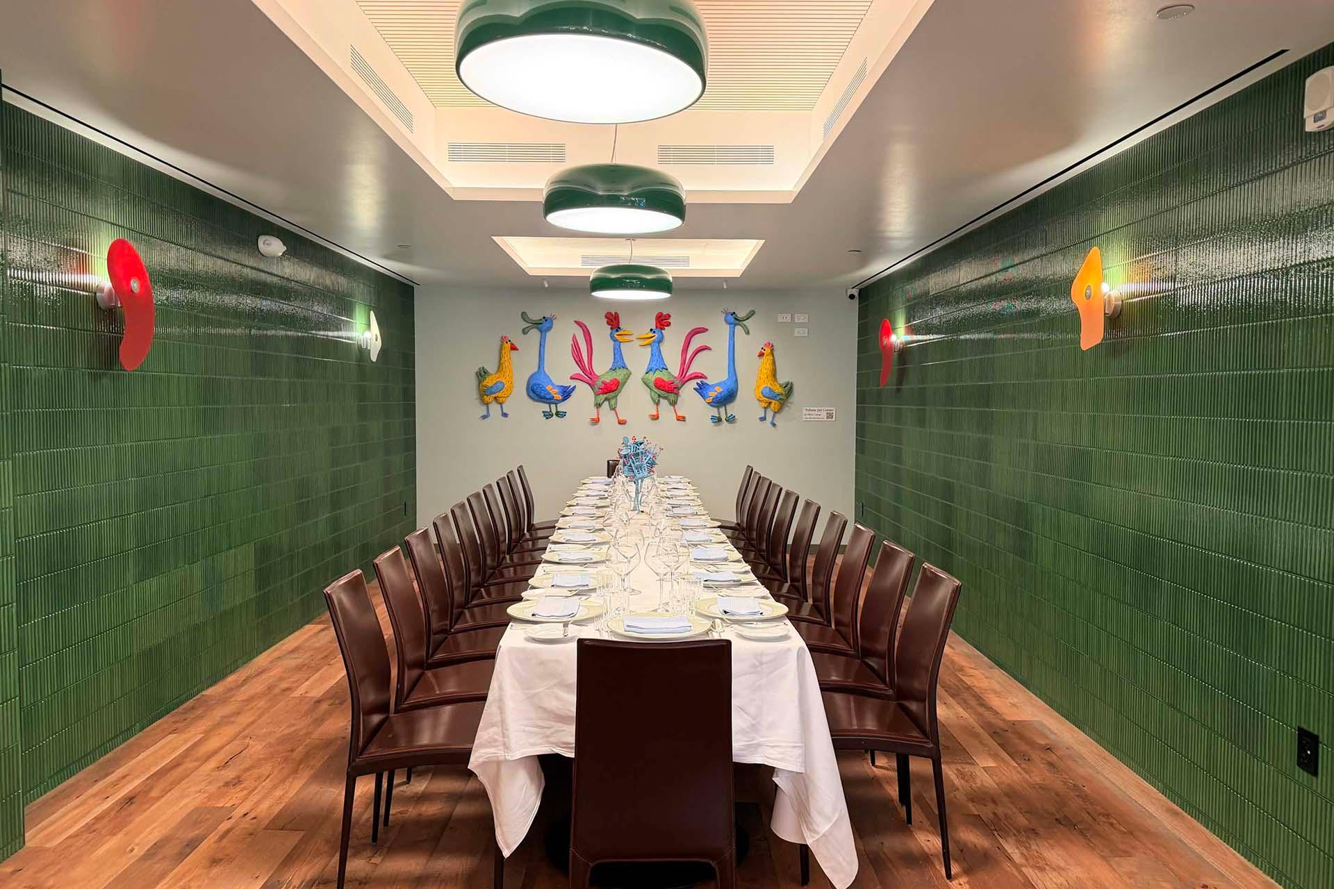 A long dining table with brown chairs sits in a room with green tiled walls, wooden floor, modern ceiling lights, and colorful rooster art on the far wall. The table is set with white cloth, plates, and glassware.