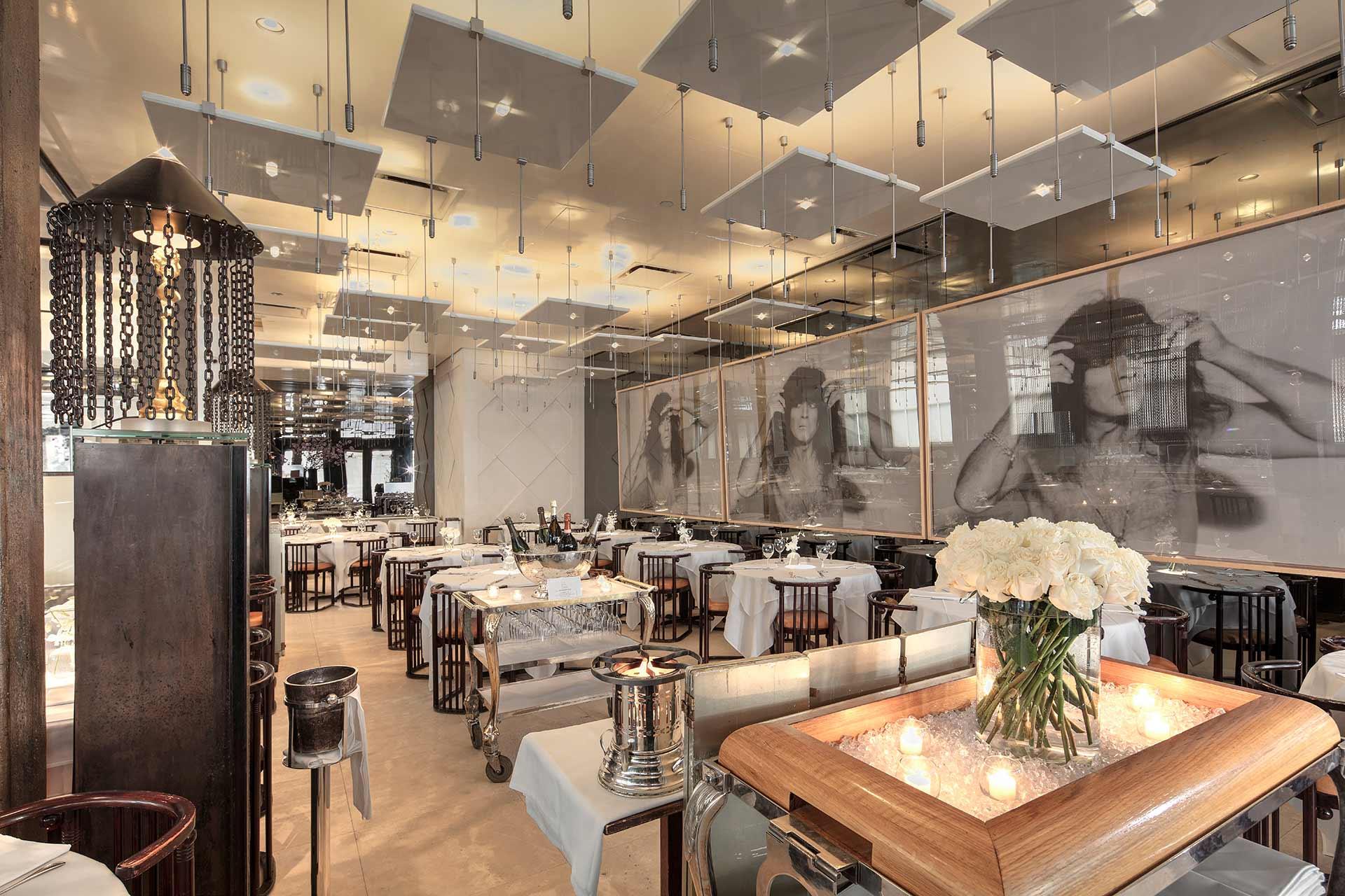 Elegant restaurant interior with neatly set tables, white tablecloths, modern lighting, large black-and-white portraits on the wall, and a centerpiece of white flowers and candles on a wooden cart in the foreground.