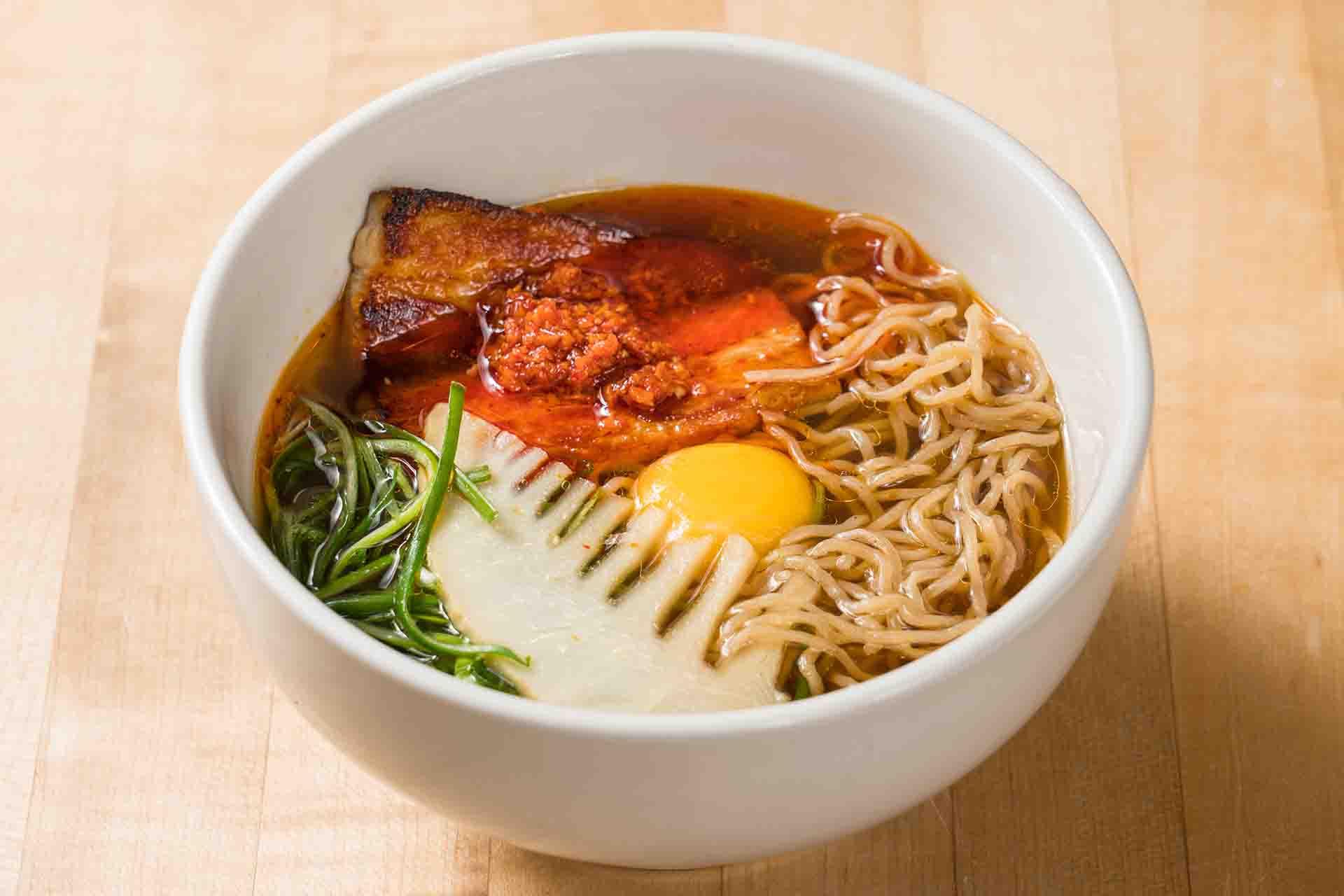 A white bowl of ramen with noodles, sliced pork, a soft-boiled egg yolk, green vegetables, bamboo shoot, and chili paste in broth sits on a light wooden surface.