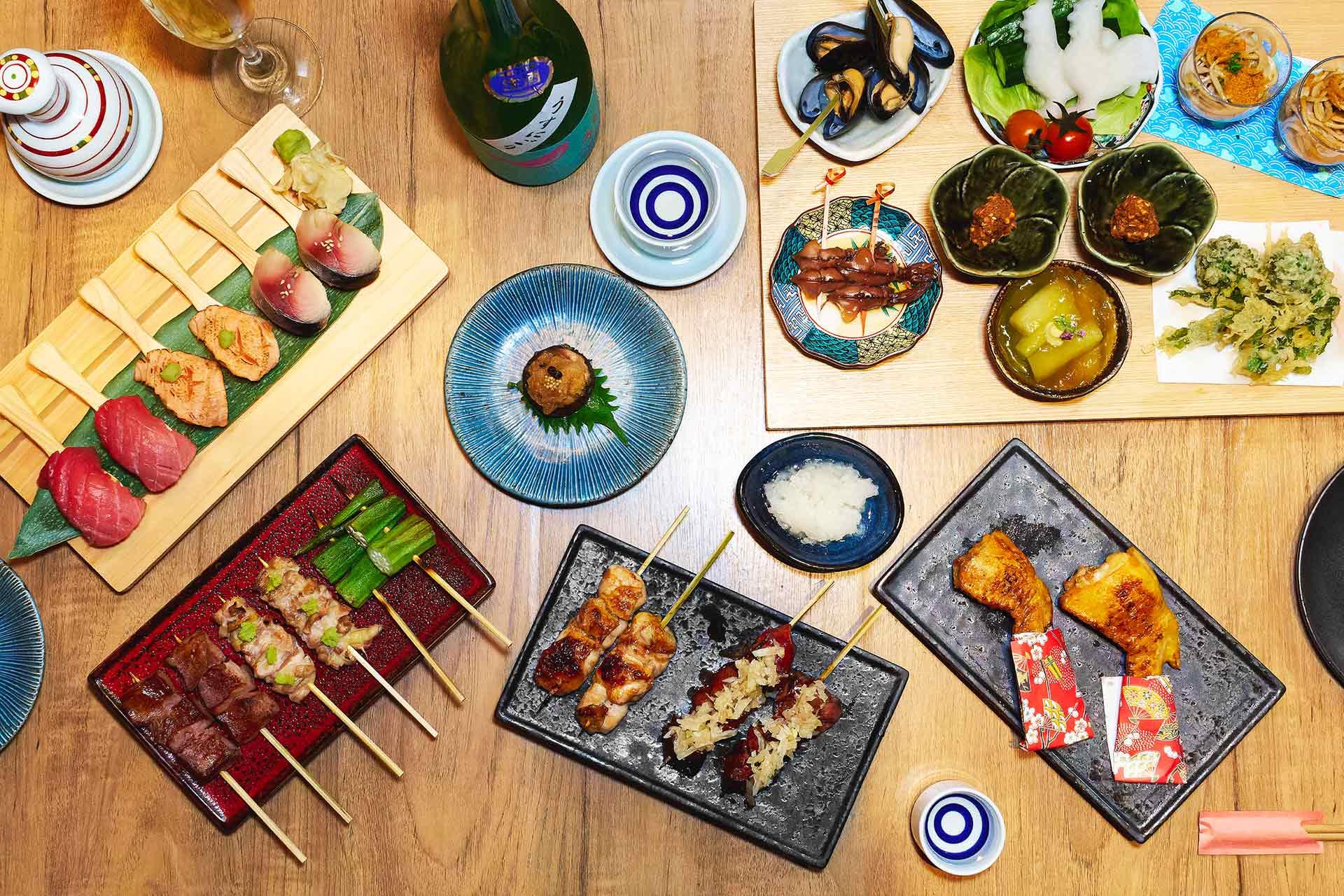 A variety of Japanese dishes, including sushi, grilled skewers, vegetables, seafood, and shared plates, are arranged on a wooden table with dipping sauces and a bottle of sake.