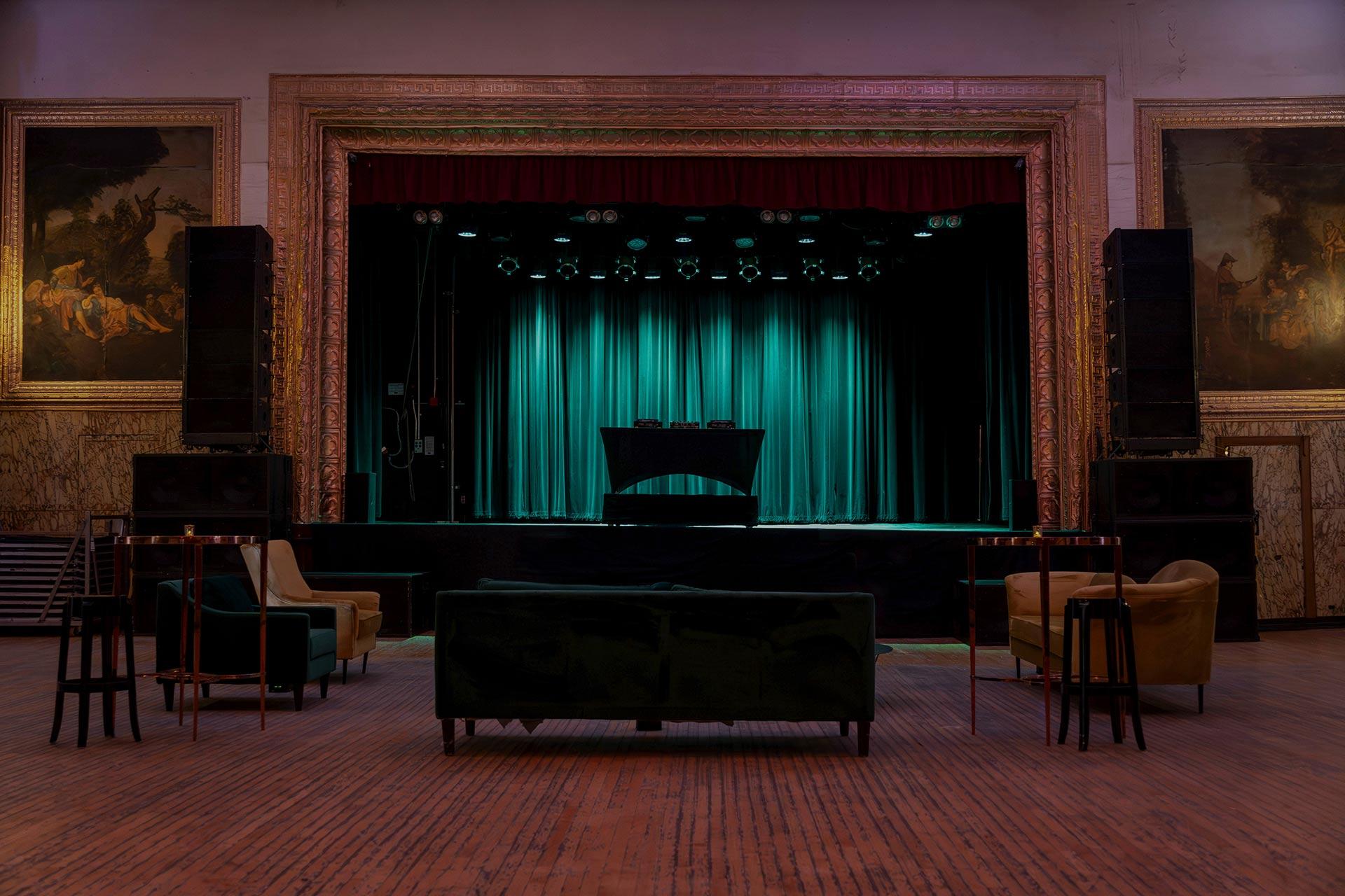 A vintage-style concert hall with green velvet curtains on stage, framed by ornate gold trim. Sofas and chairs are arranged lounge-style on the wooden floor, with large paintings on the walls.