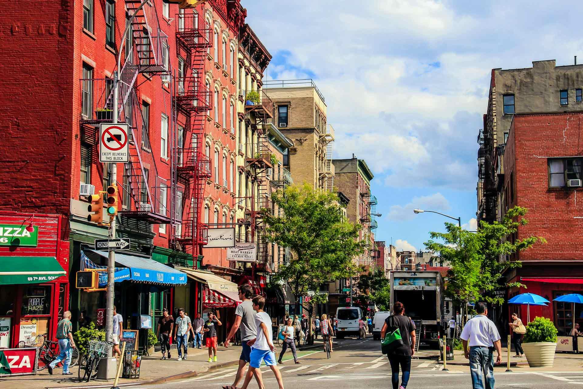 A bustling city street corner with pedestrians, red brick buildings, and a variety of storefronts. A delivery truck and several cars are on the road. Trees line the street, and a blue sky with clouds is above.