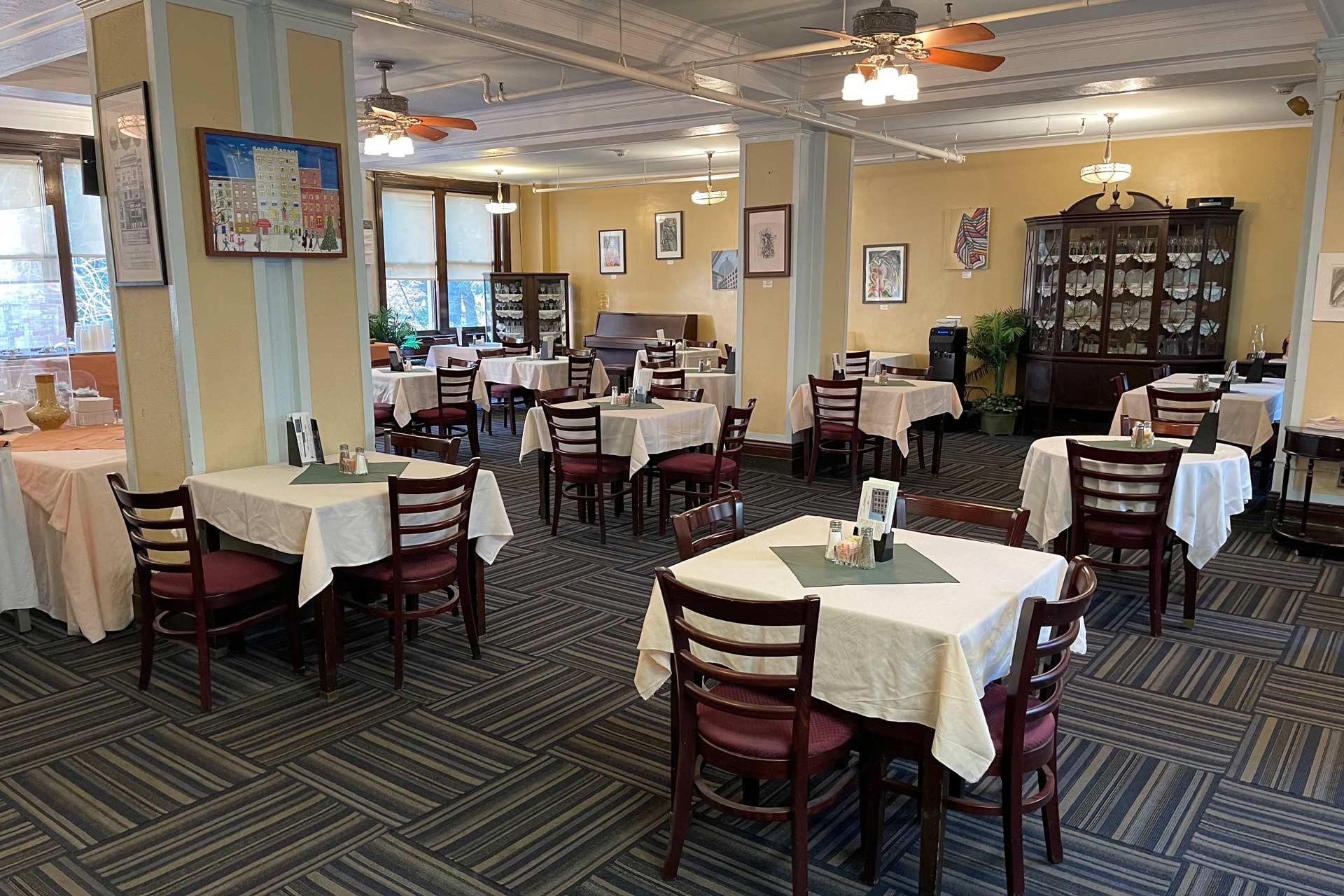 A spacious, well-lit restaurant dining room with several tables covered in cream-colored tablecloths, wooden chairs, striped carpet, framed art on yellow walls, ceiling fans, and a china cabinet in the corner.
