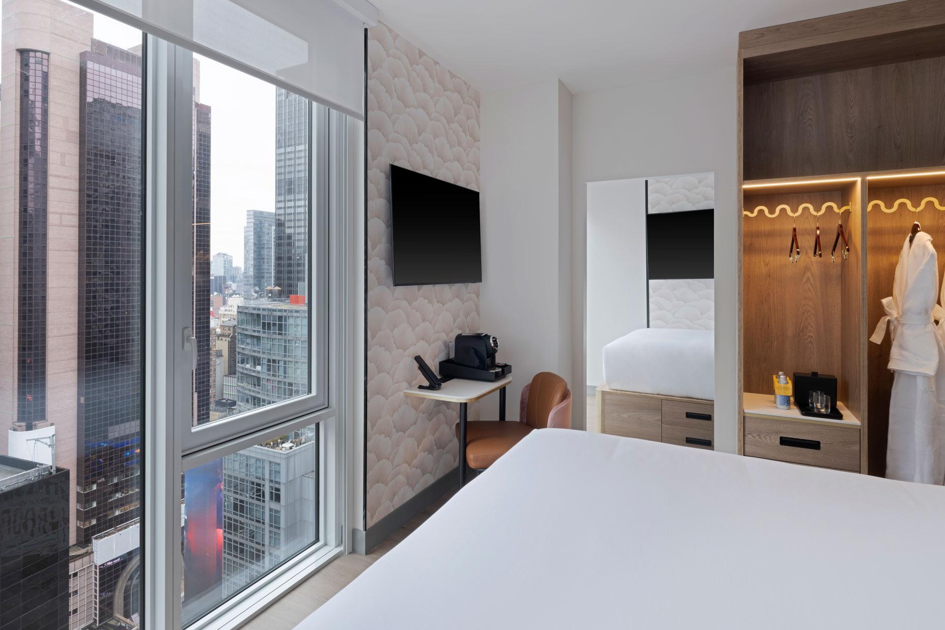 A modern hotel room with a large bed, wall-mounted TV, desk, open closet, and city view through floor-to-ceiling windows. A robe hangs in the closet, and tall buildings are visible outside.