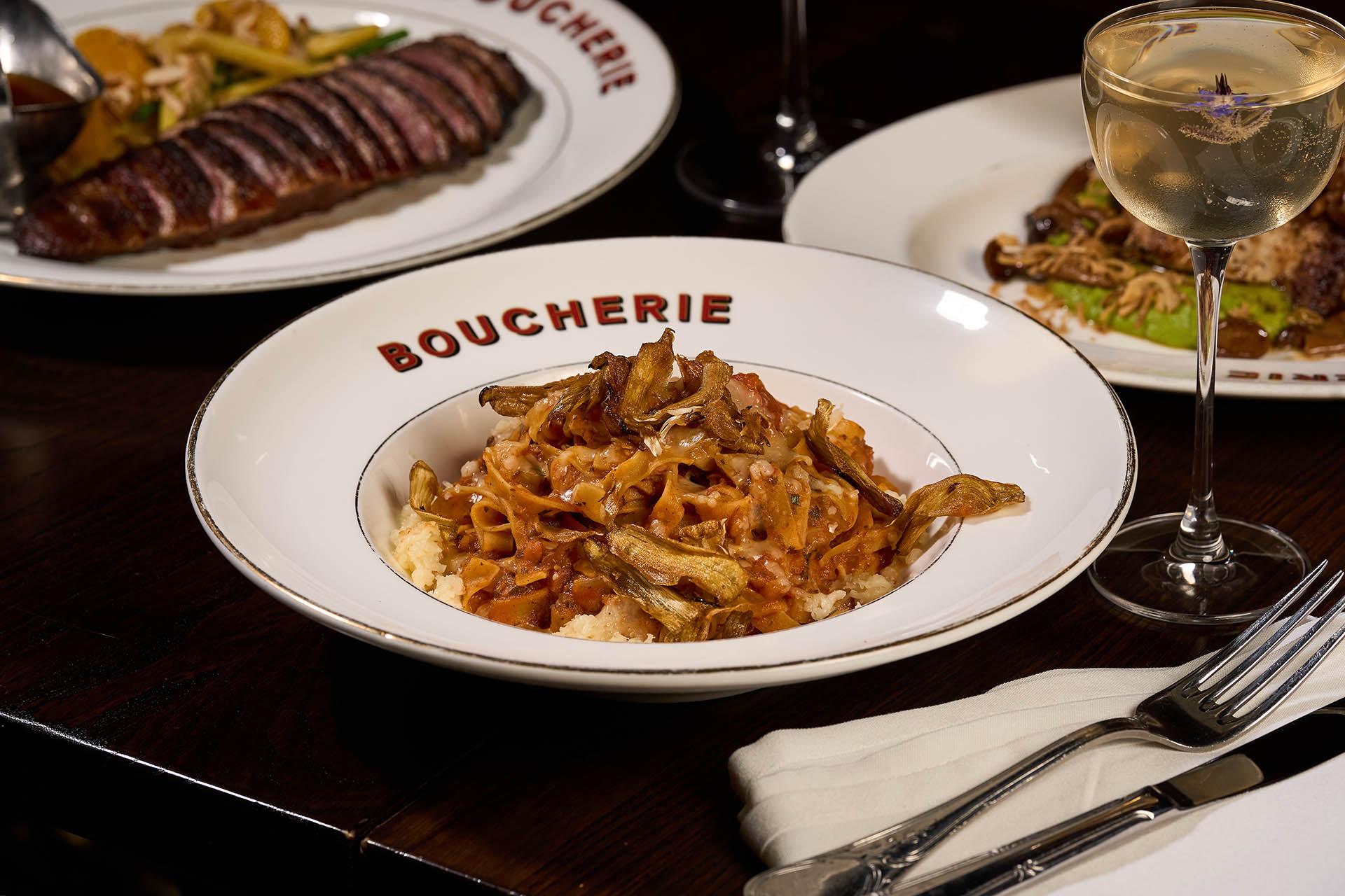 A bowl of pasta topped with mushrooms sits on a “BOUCHERIE” plate, accompanied by a glass of white wine and plates of steak and meat in the background on a dark wooden table.