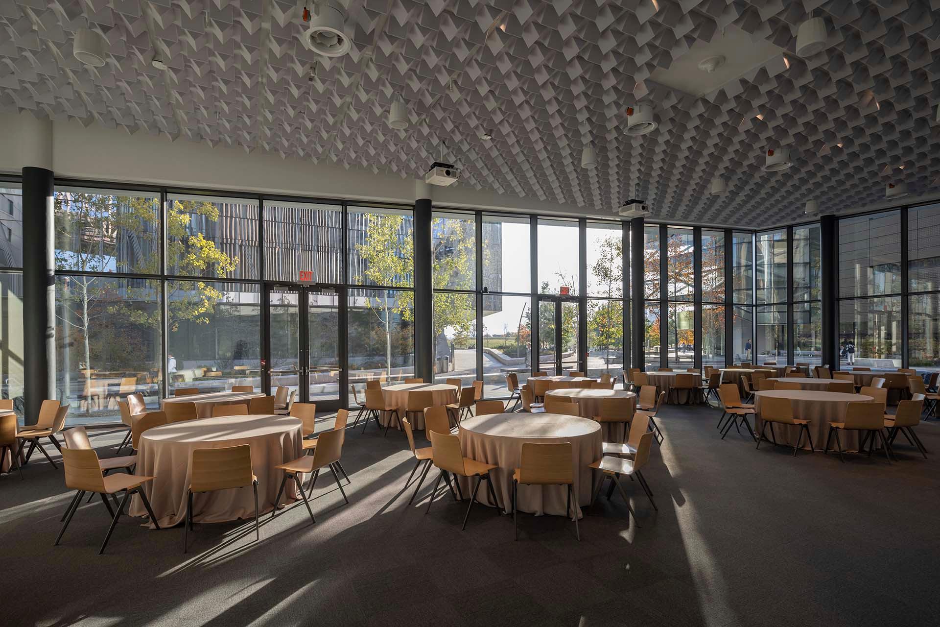 A modern event space with round tables covered in beige cloths, wooden chairs, large floor-to-ceiling windows, and geometric ceiling panels. Sunlight streams in, creating shadows on the floor.