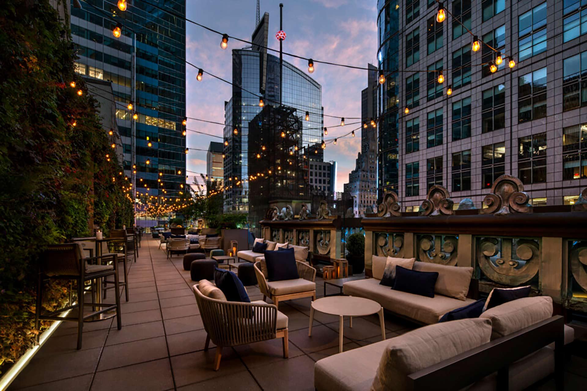 Outdoor rooftop lounge at dusk, surrounded by tall modern buildings, with string lights hanging overhead, cozy sofas and chairs, greenery on one wall, and a relaxed, inviting atmosphere.
