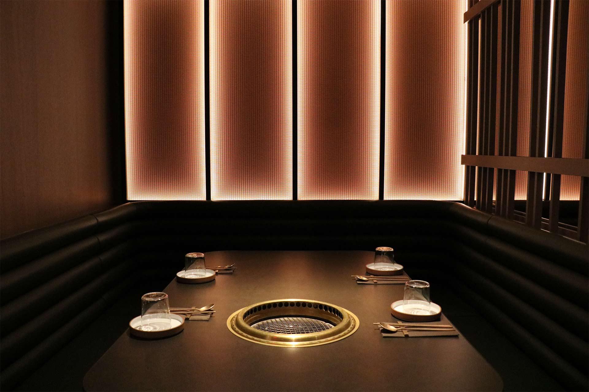 A modern restaurant booth with a built-in grill at the center of the table, set with four plates, glasses, and utensils. Soft, warm lighting illuminates the minimalist, cozy setting.