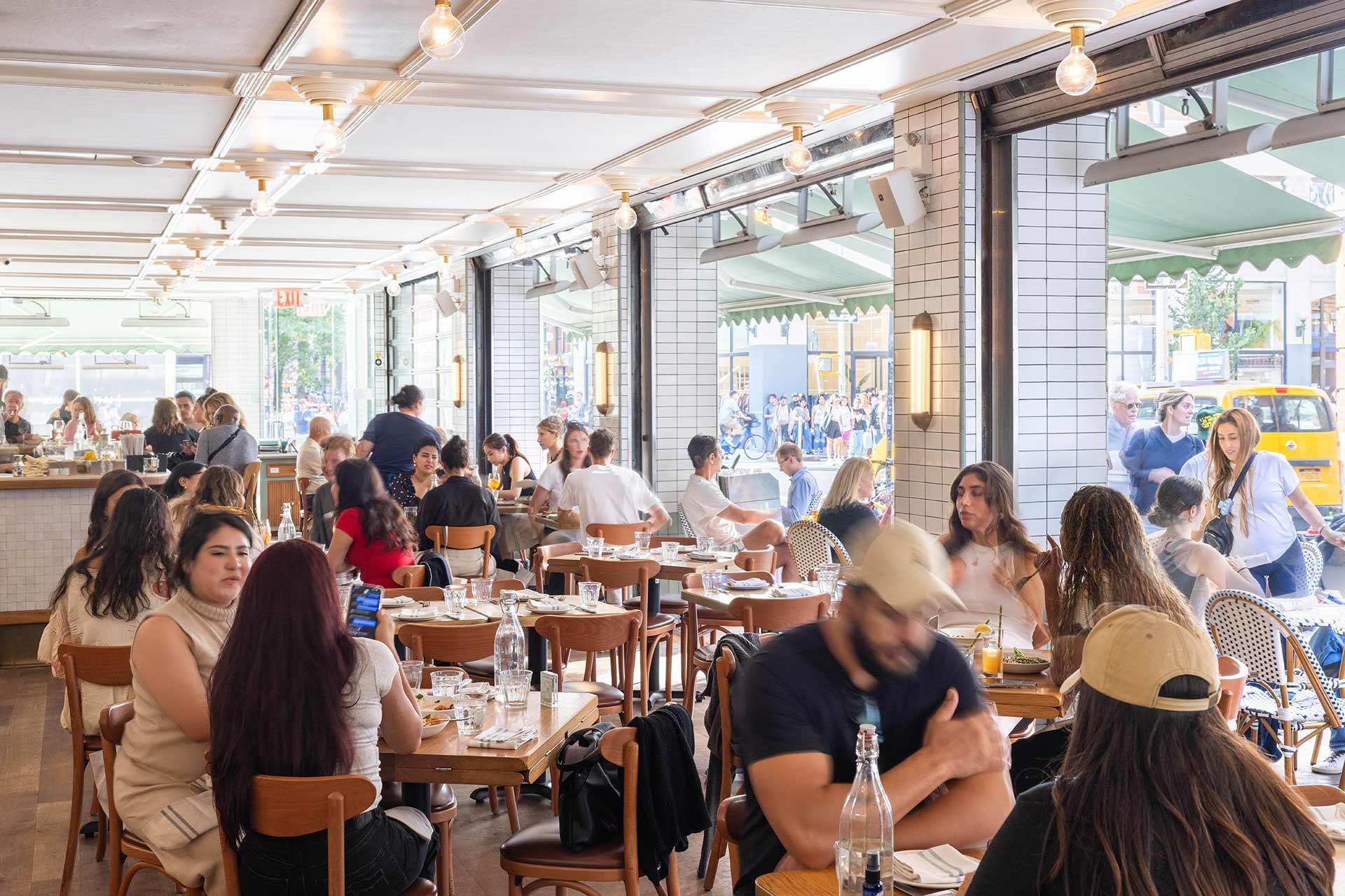 A bright, busy restaurant filled with people dining at wooden tables, large windows letting in natural light, and outdoor seating visible through the open doors. The atmosphere is lively and modern.