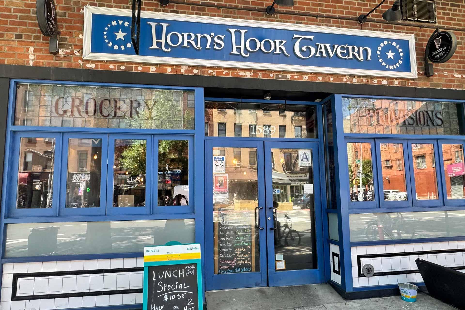 Exterior of Horn's Hook Tavern, a blue-fronted bar and restaurant with large windows, a sidewalk chalkboard sign advertising lunch specials, and reflections of buildings across the street.