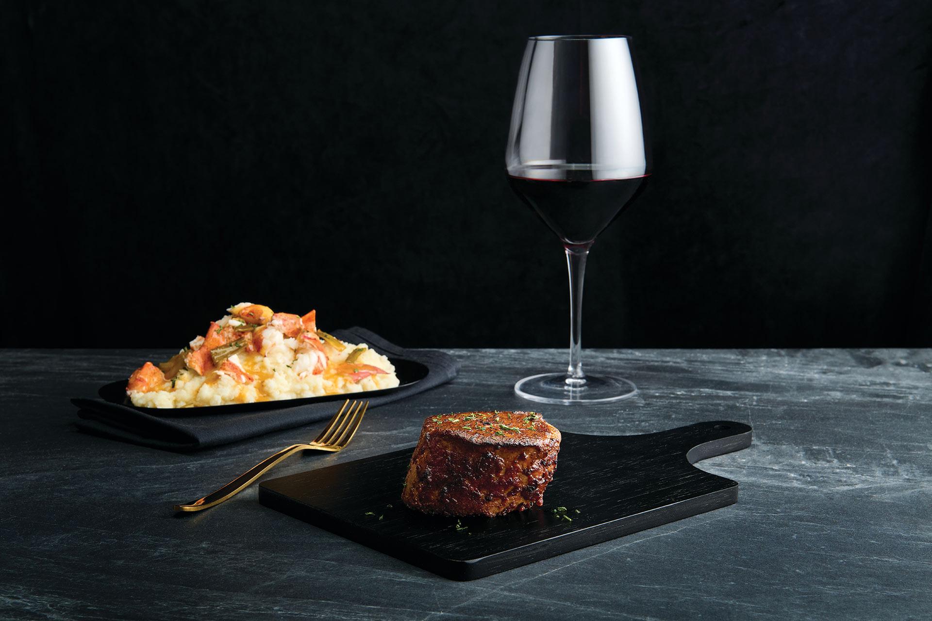 A glass of red wine, a serving of mashed potatoes with sauce and herbs on a black plate, and a cooked filet mignon steak on a black wooden board, all arranged on a dark stone surface.