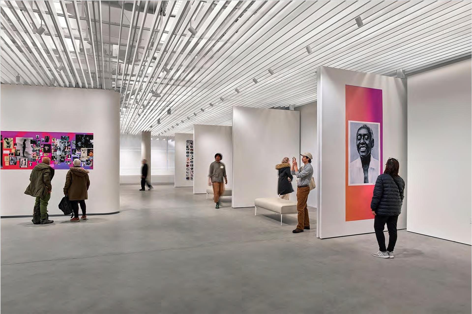 People view artwork displayed on large, white walls in a modern, spacious art gallery with high ceilings and ample lighting. Some visitors stand, while others sit on benches, observing various colorful and monochrome pieces.