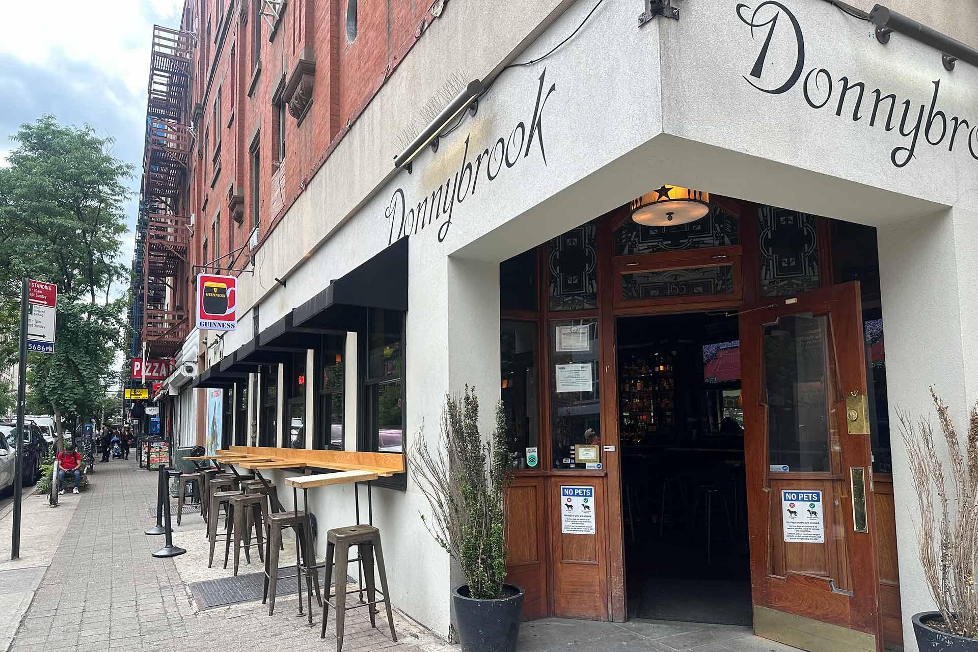 A street view of Donnybrook bar with open wooden doors, outdoor seating with stools along a counter, and a tree-lined sidewalk on a cloudy day. Signs and neighboring buildings are visible.