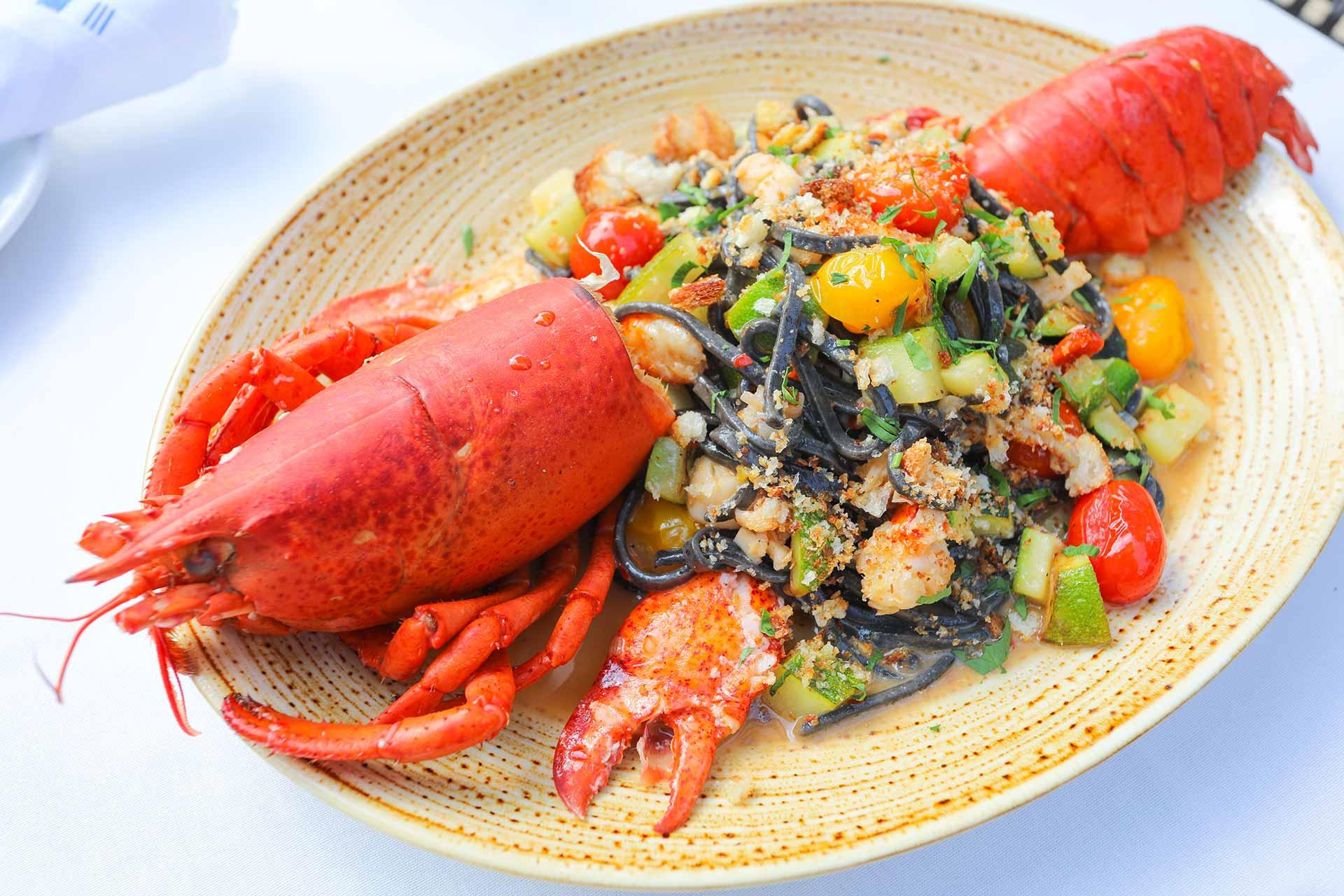 A plate of black squid ink pasta topped with diced vegetables and herbs, garnished with a cooked lobster split in half, served on a beige dish on a white tablecloth.