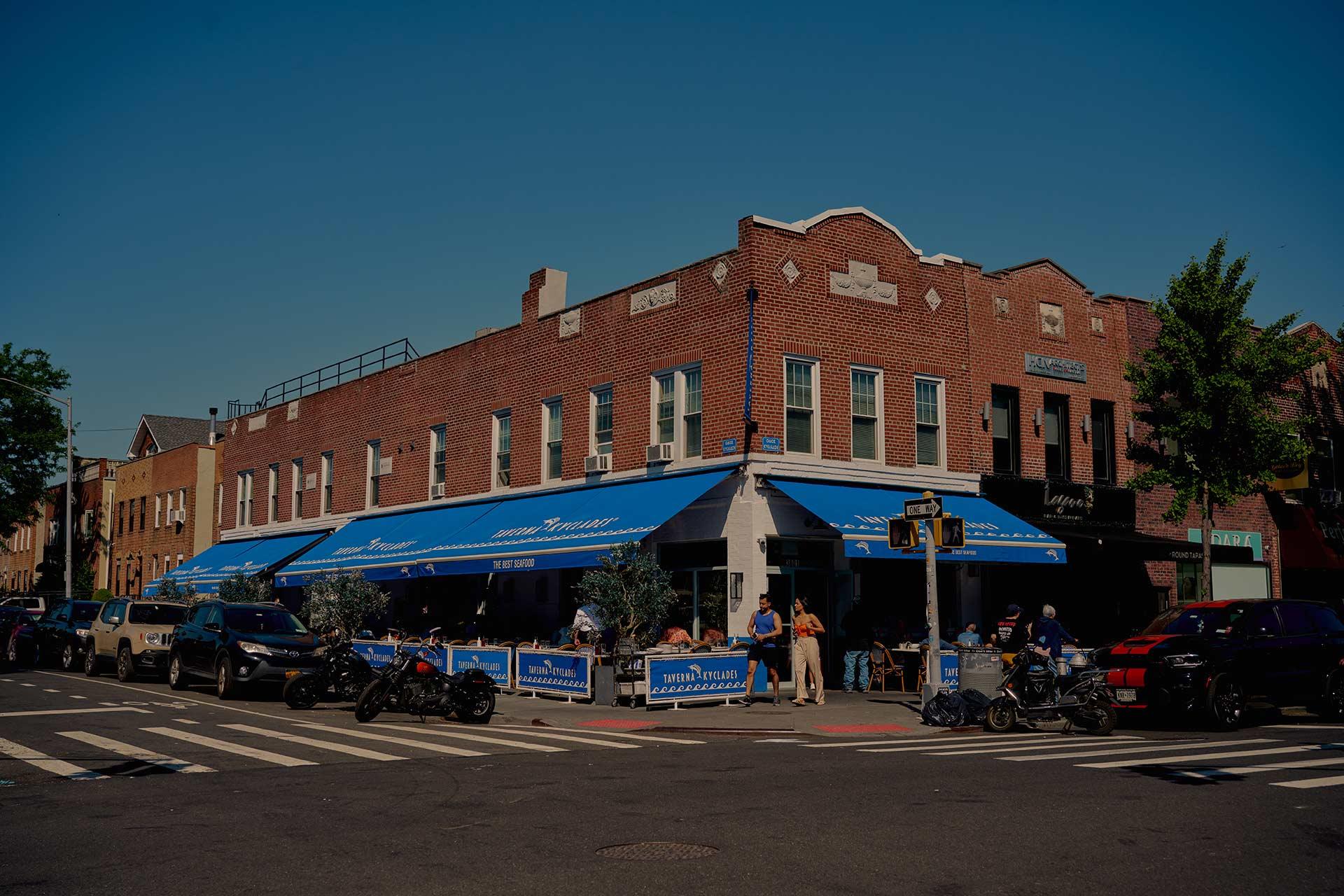 A corner brick building with blue awnings houses a restaurant with outdoor seating. People dine at tables on the sidewalk; cars and motorcycles are parked along the street under a clear blue sky.