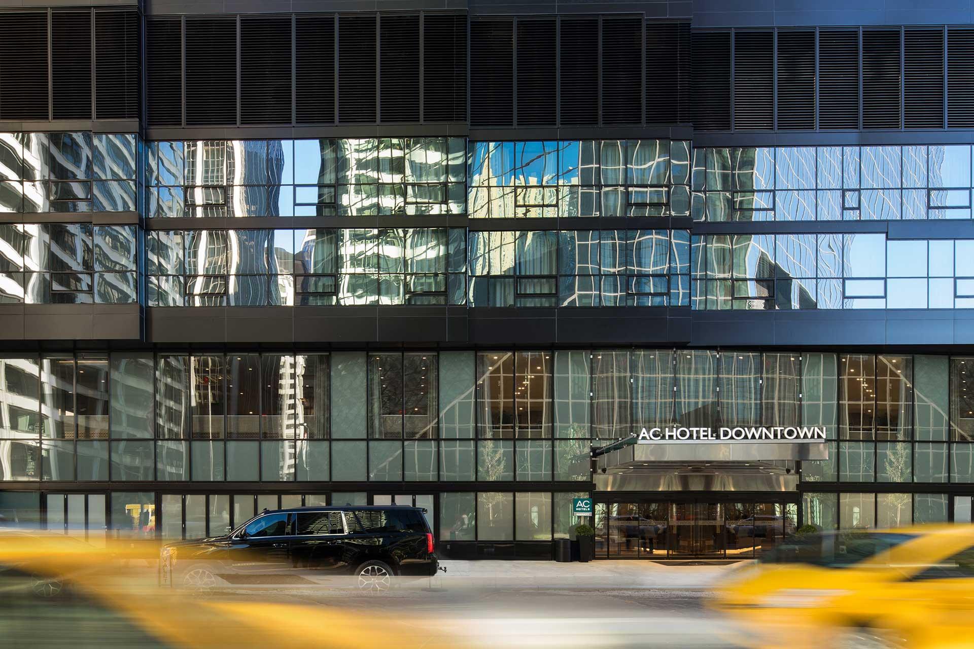 A modern glass building with the sign "AC Hotel Downtown" above the entrance; city skyscrapers are reflected in the windows, and yellow cars blur past on the street in front.