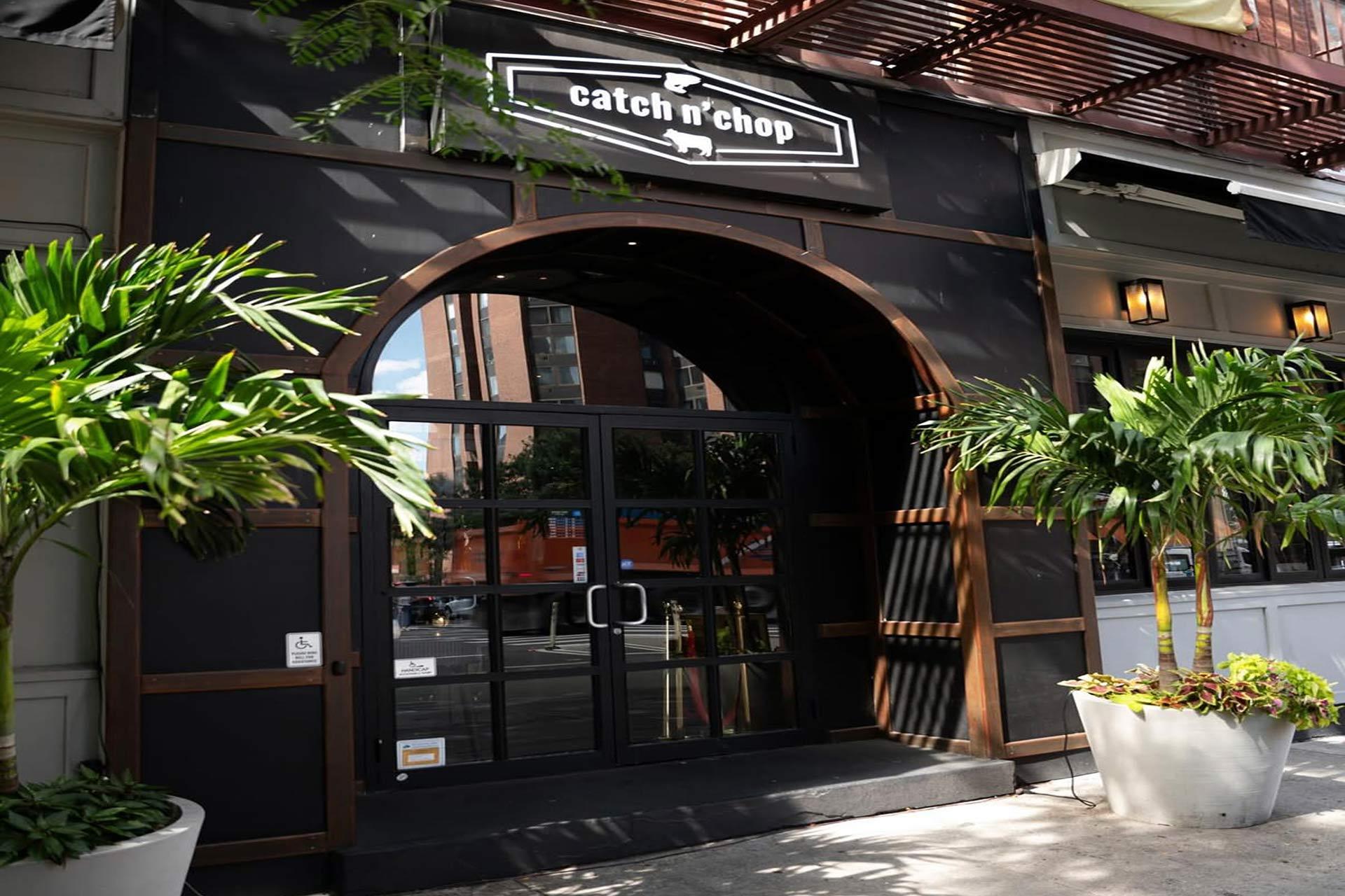 The entrance to a restaurant called "catch n’ chop," featuring a large arched doorway, double glass doors, and potted palm trees on either side, with city buildings reflected in the glass.