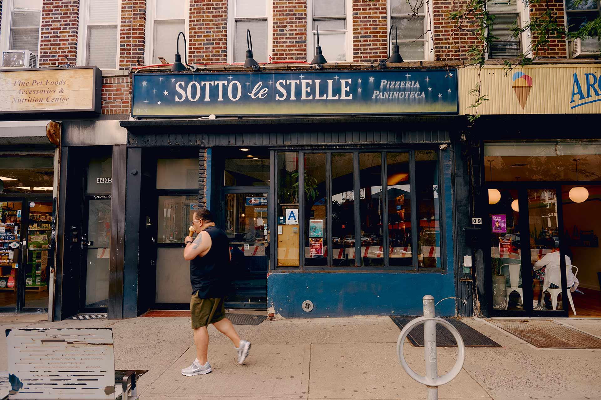 A person walks past the storefront of “Sotto le Stelle,” a pizzeria and paninoteca on a city street, with brick buildings and other shops visible on either side.