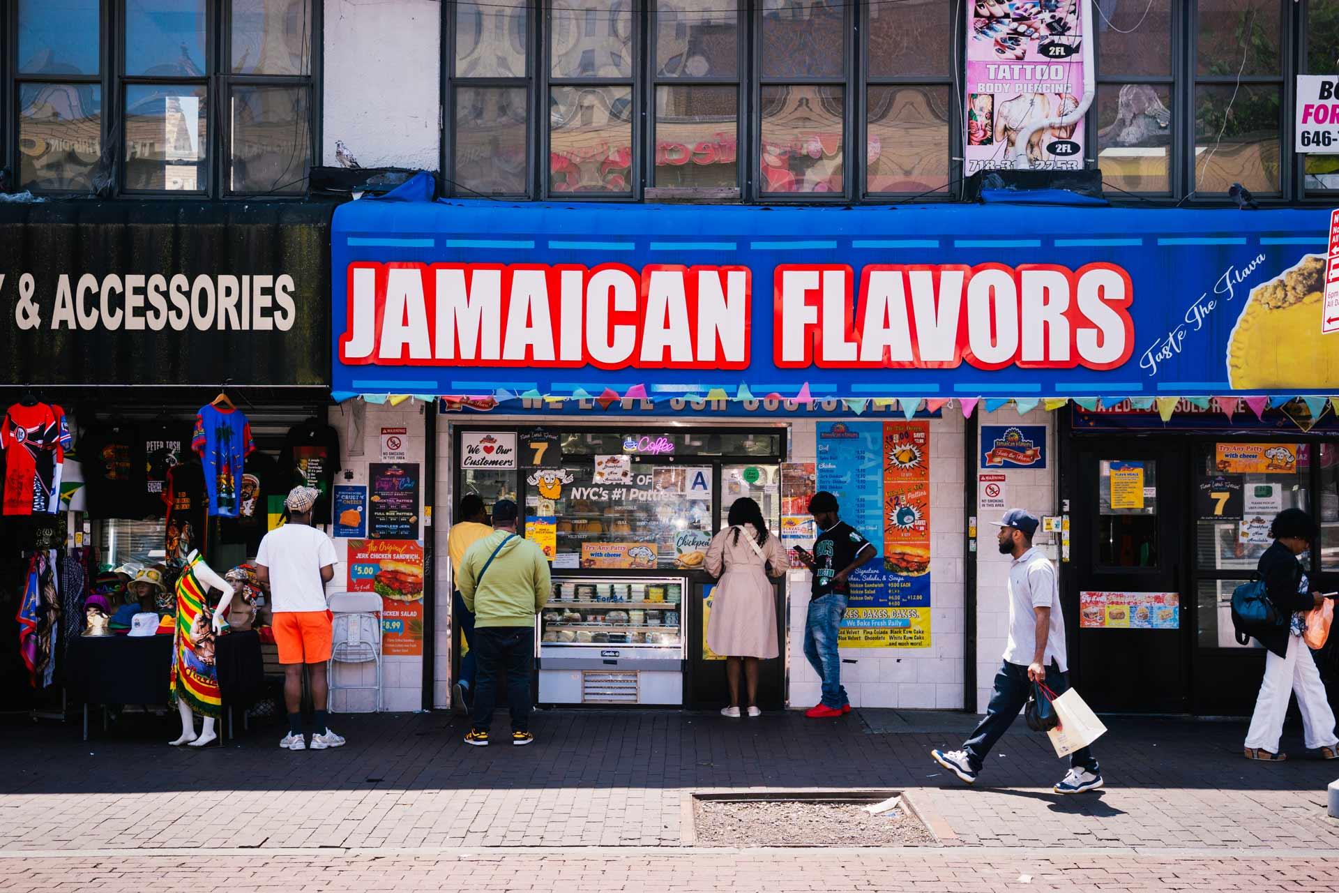 People stand in front of a store called "Jamaican Flavors" with a bright blue and red sign. The sidewalk is busy with pedestrians, and colorful posters and clothing are displayed outside nearby shops.
