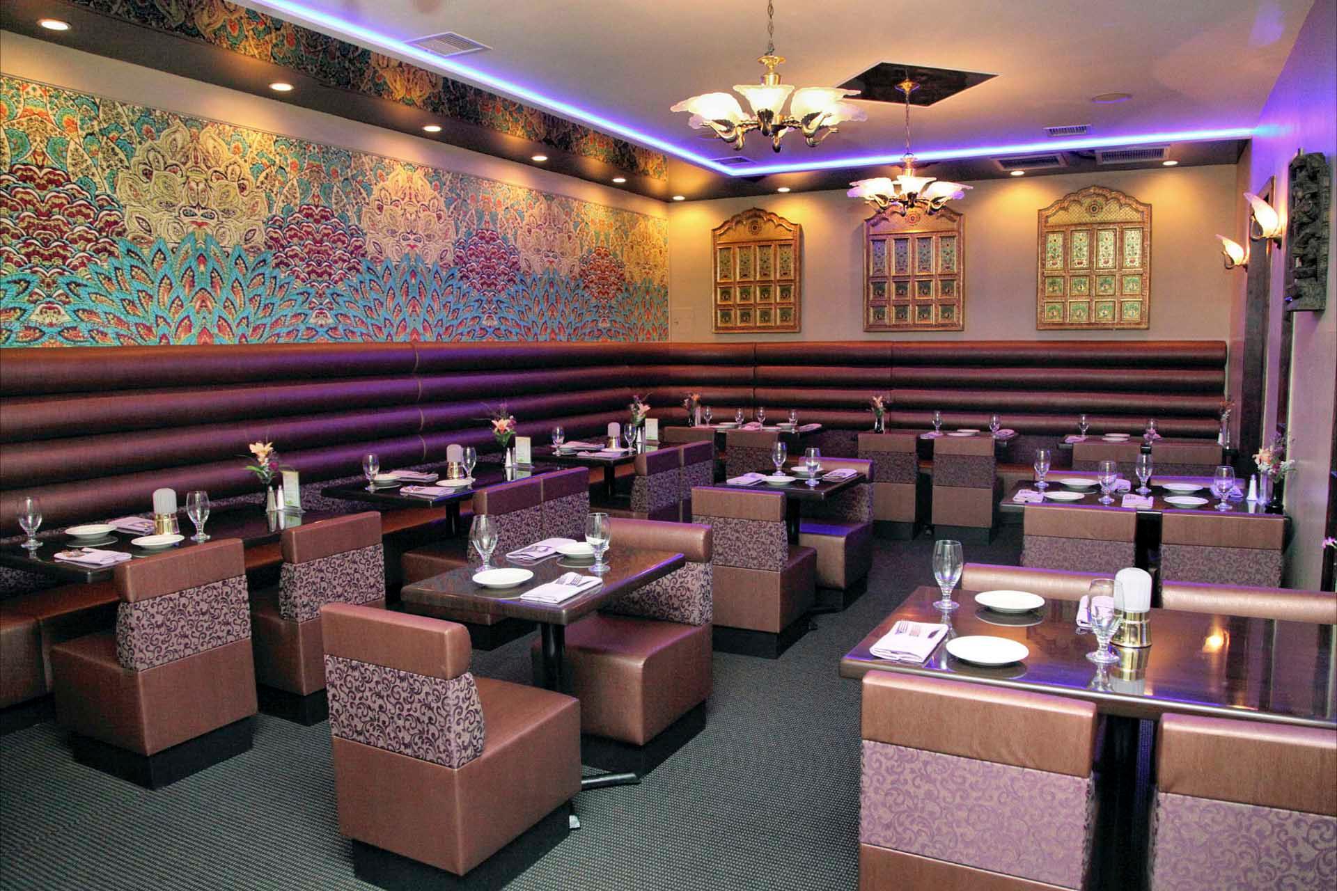 An elegant restaurant interior with neatly arranged tables and cushioned chairs. The walls are adorned with intricate patterns in vibrant colors, and stylish chandeliers hang from the ceiling, casting a warm glow over the dining area.