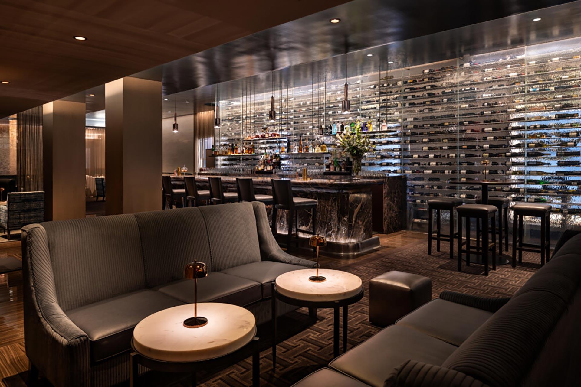 A modern, dimly lit lounge with plush gray sofas, round marble tables, and a stylish bar in the background. The bar features a wall of wine bottles and elegant lighting, creating a cozy, upscale atmosphere.