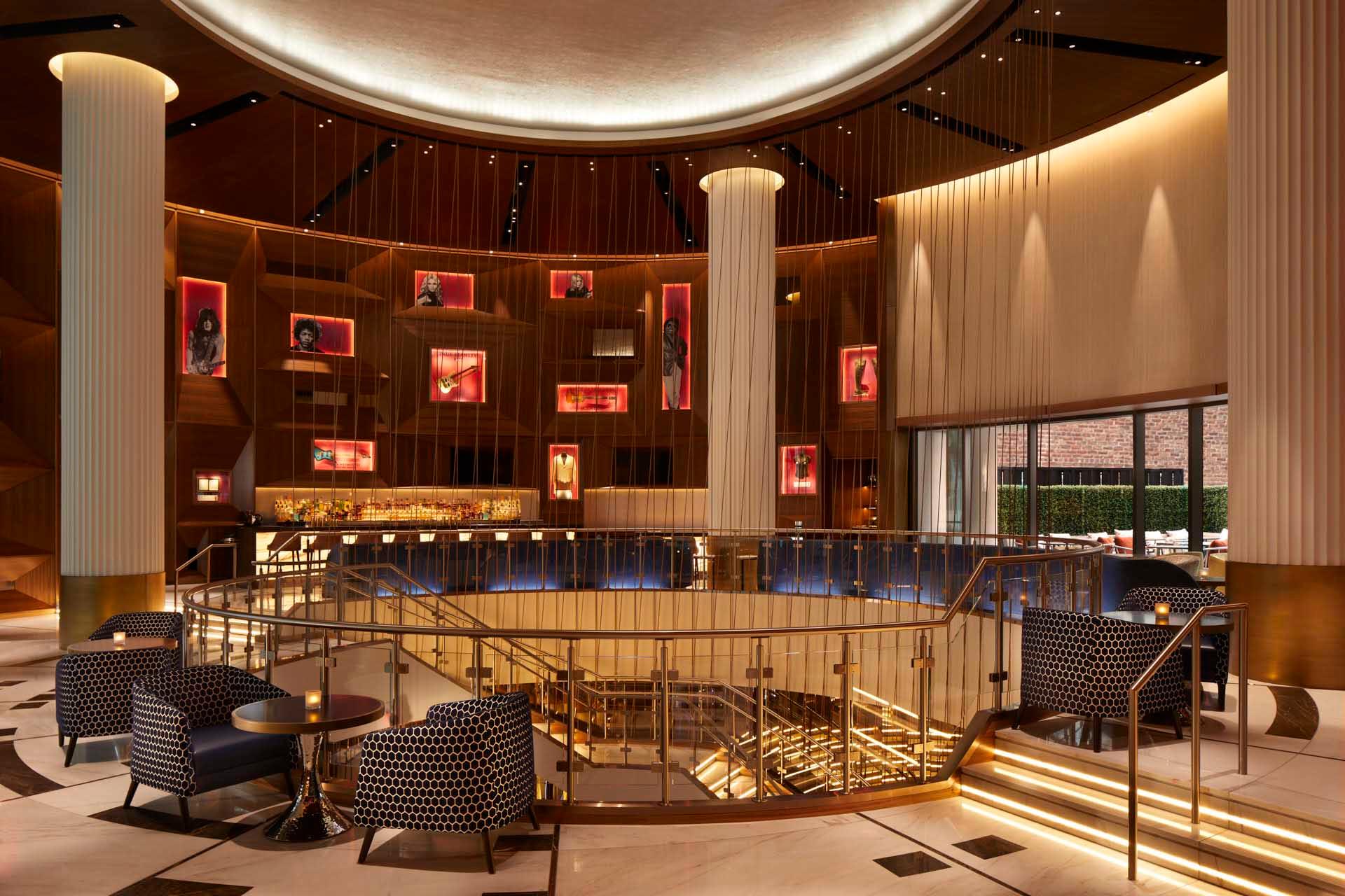 Luxurious hotel lobby with a circular design, featuring a central staircase, large columns, and ambient lighting. Modern seating with patterned chairs surrounds the area. Artwork is displayed on a wooden wall with illuminated frames.