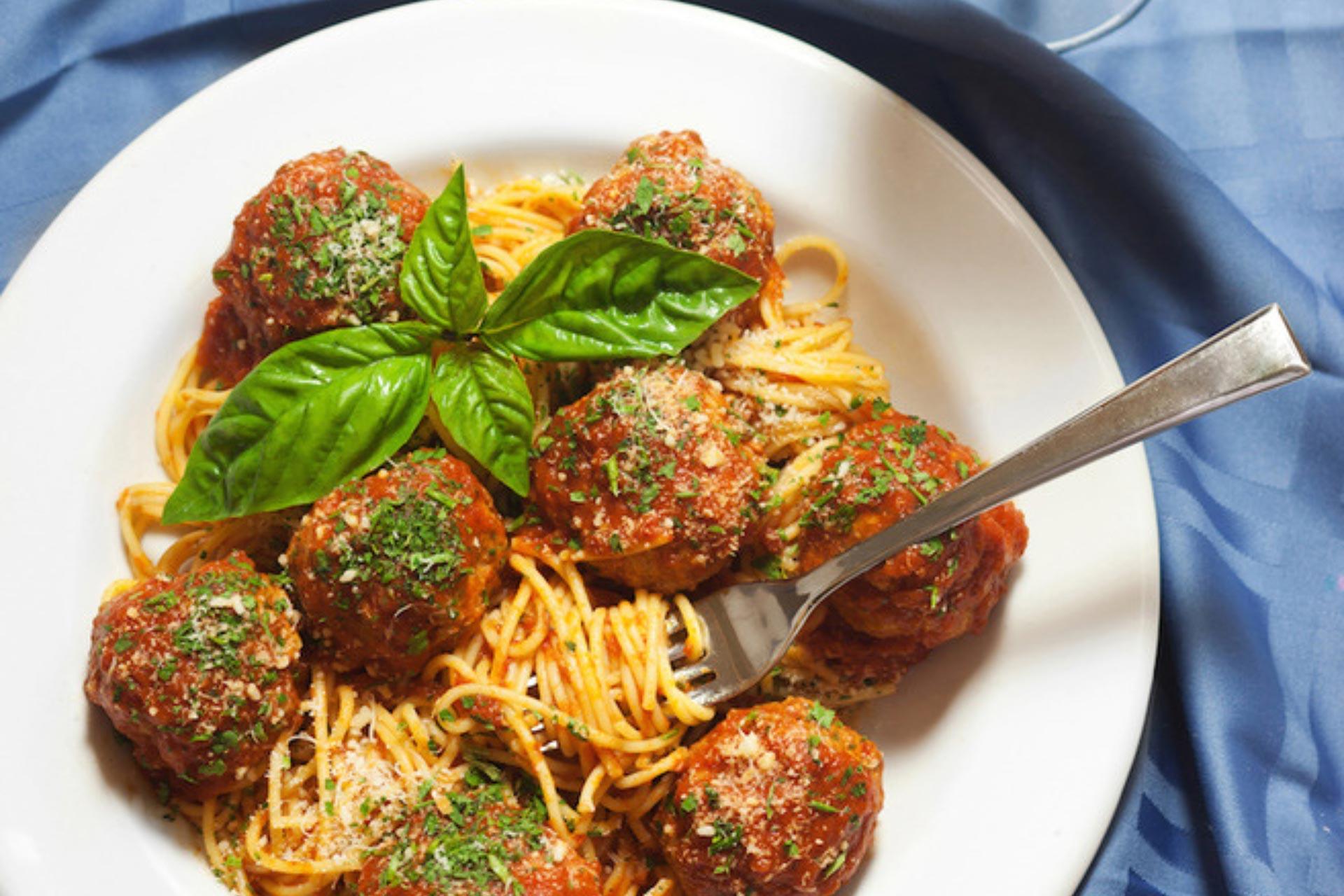 A plate of spaghetti topped with marinara sauce, meatballs, grated cheese, chopped herbs, and a garnish of fresh basil, with a fork resting on the side. The plate sits on a blue tablecloth.
