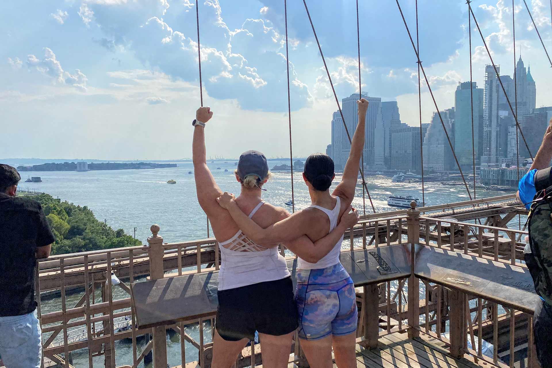 Two people in athletic wear stand arm-in-arm on the Brooklyn Bridge, raising their fists in celebration while overlooking the East River and the New York City skyline on a sunny day.