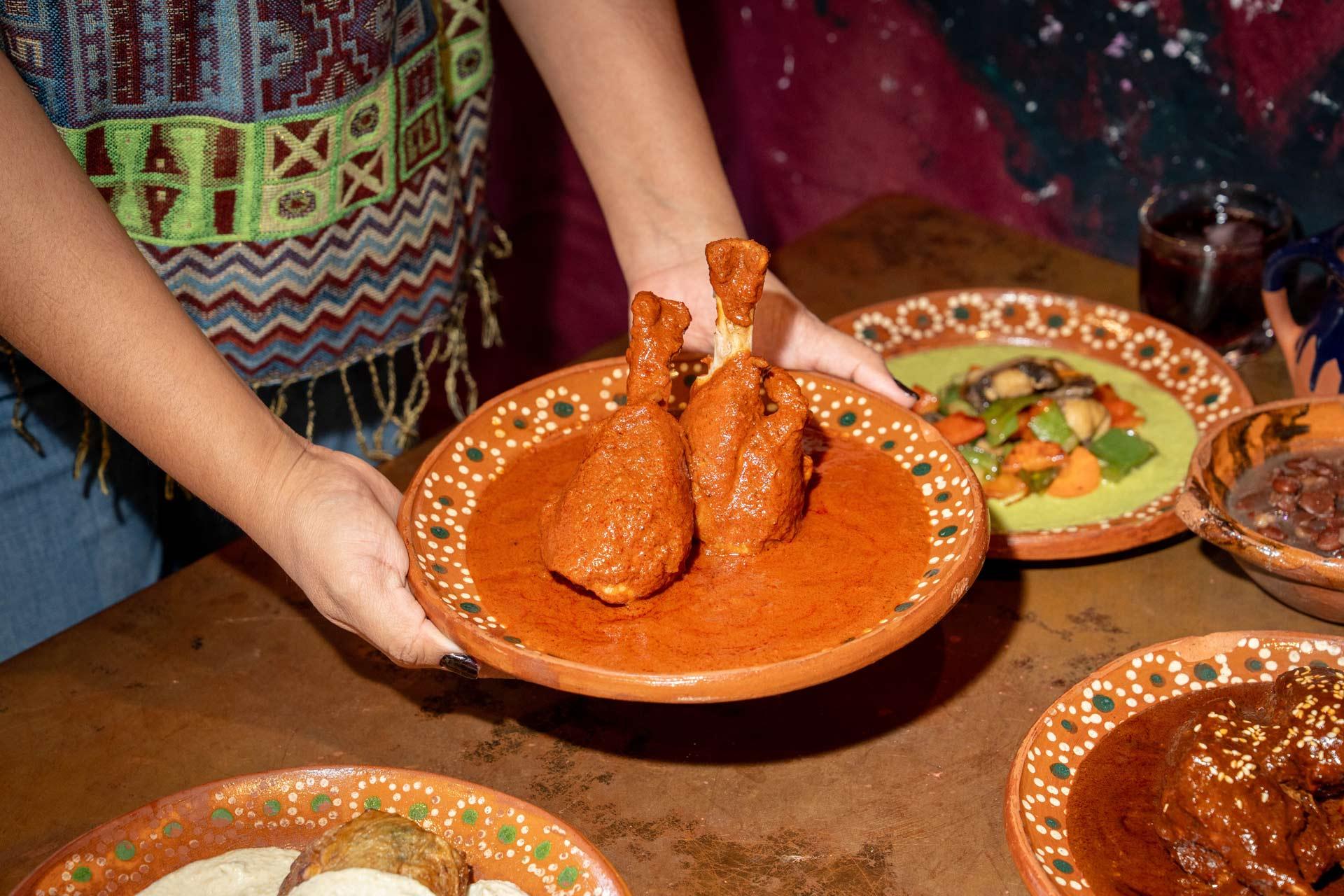 A person holds a clay plate with two chicken drumsticks covered in mole sauce. Other colorful Mexican dishes on clay plates are on the wooden table nearby. The person wears a patterned shirt.