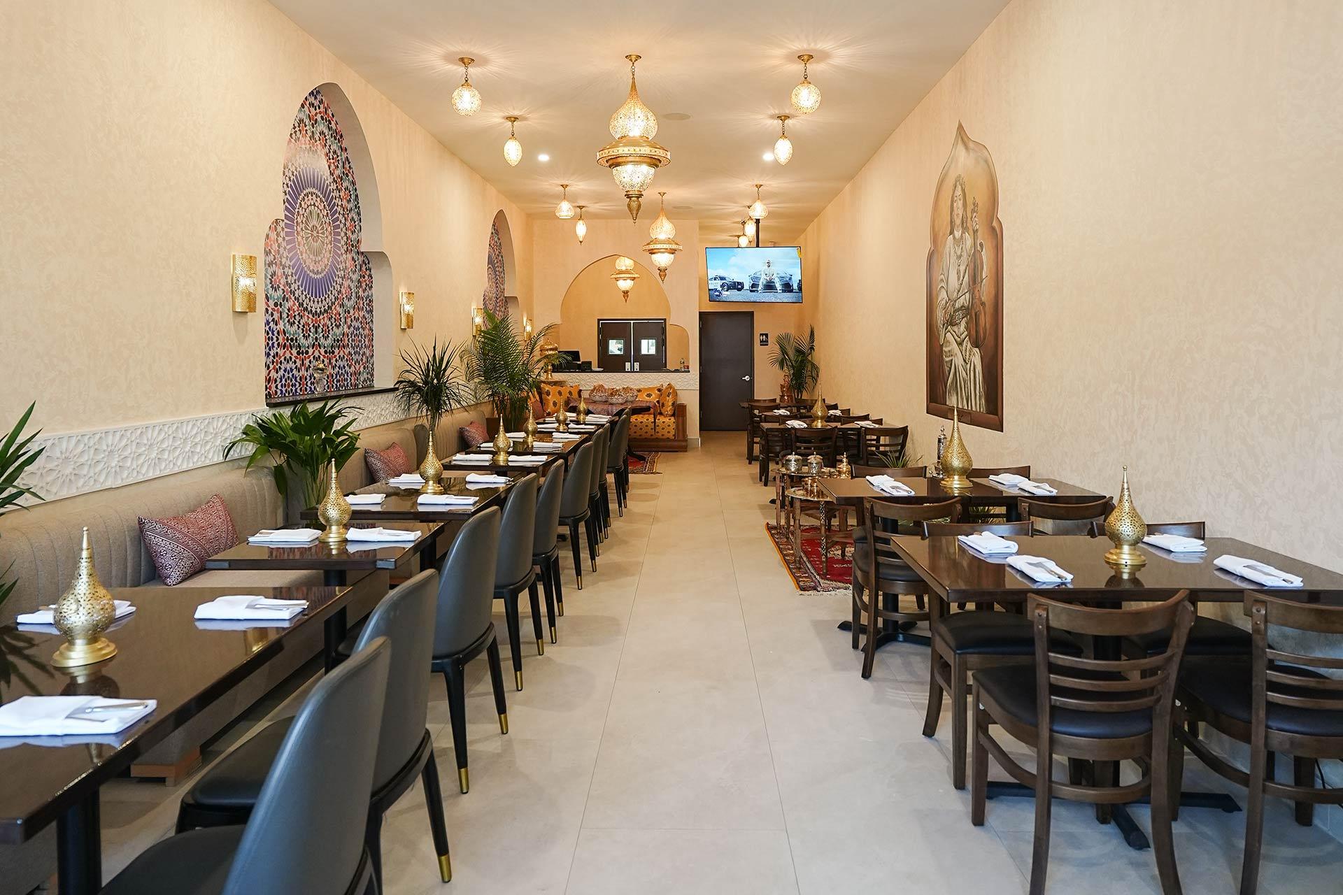 A cozy restaurant with beige walls, decorative chandeliers, wall art, cushioned seating on one side, and dark wooden tables and chairs set for dining. Plants and pillows add a warm, inviting atmosphere.