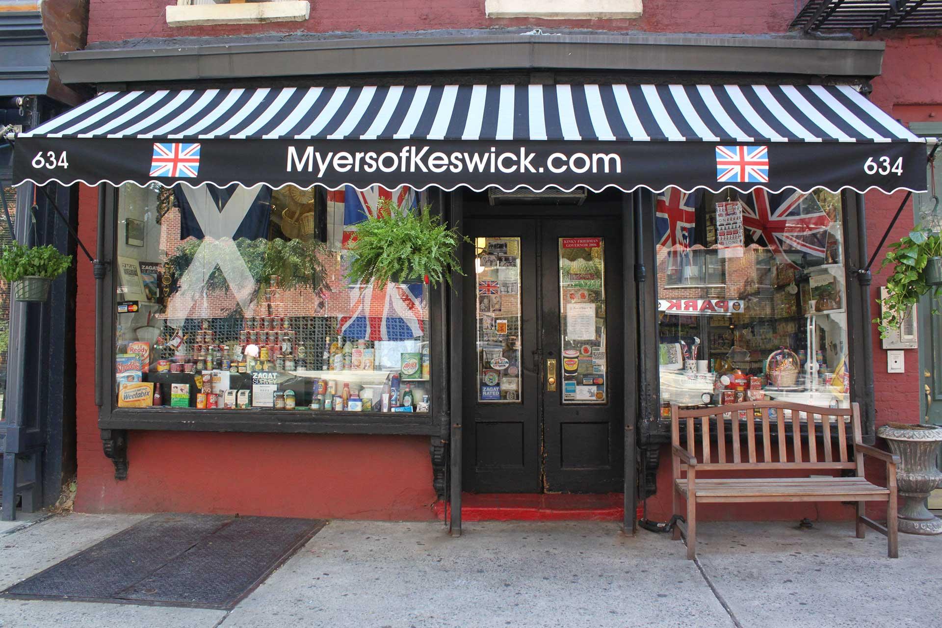 A storefront with a black-and-white striped awning that reads "MyersofKeswick.com" and features British flags. The shop window displays various goods, and a wooden bench sits outside by the entrance.