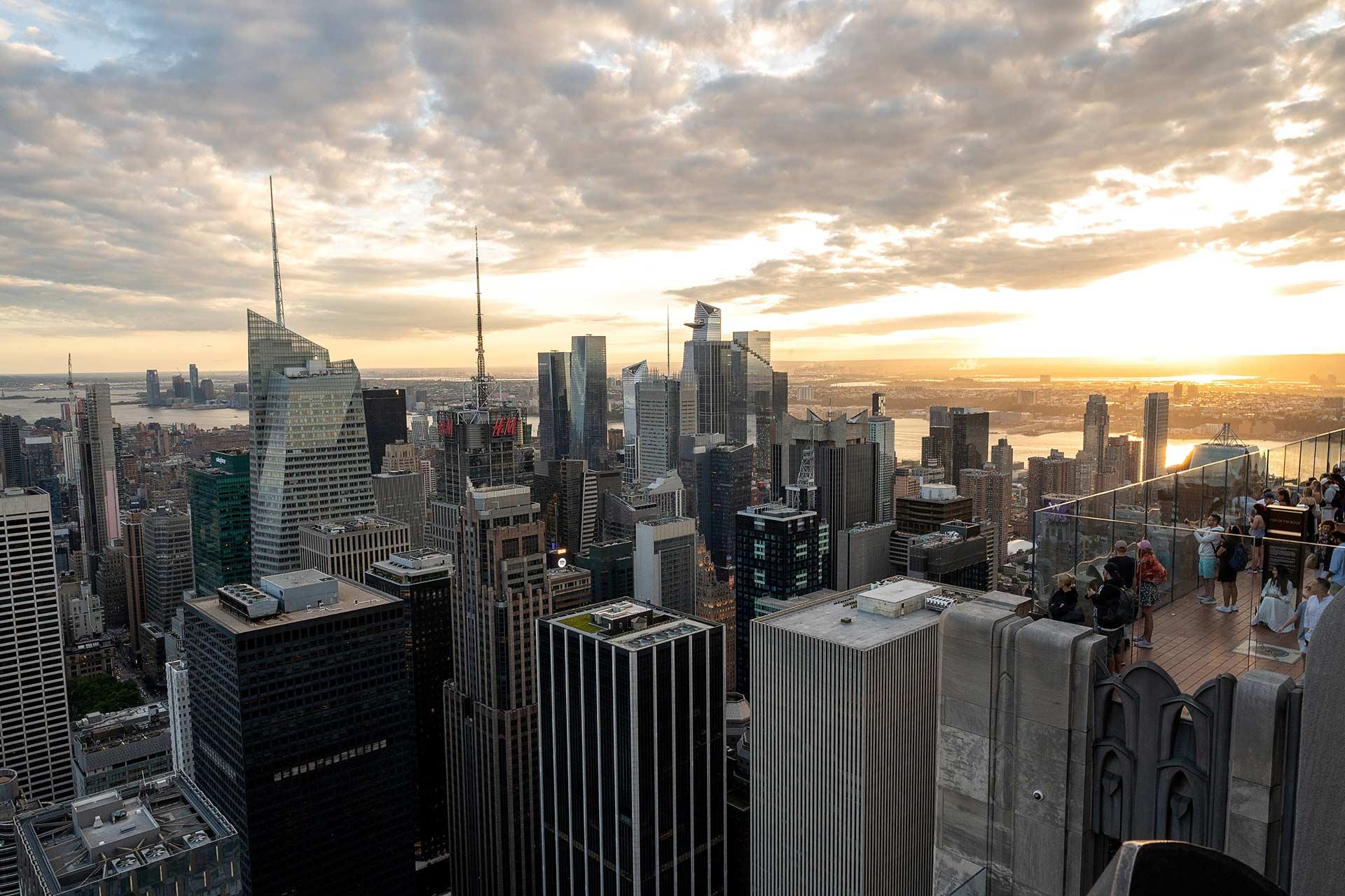 A view of the New York City skyline at sunset, with tall skyscrapers under a partly cloudy sky and people standing on an observation deck in the foreground.