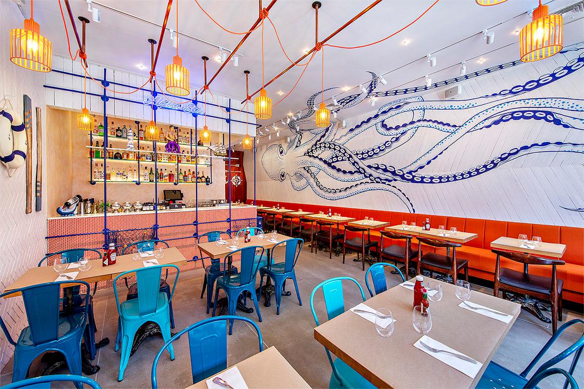 A modern restaurant interior featuring teal metal chairs, orange cushioned benches, and hanging woven light fixtures. The walls showcase a blue octopus mural. The bar at the back is stocked with bottles and has a hanging light system with red wires.