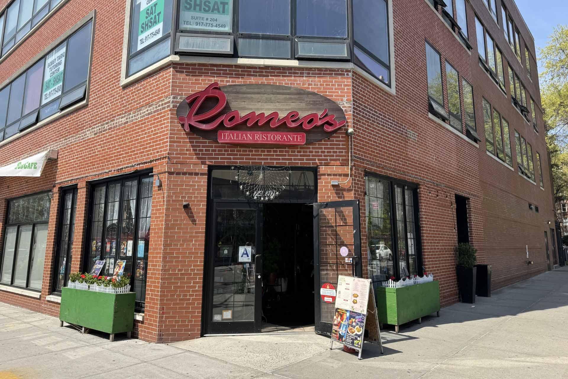 Street view of Romeo’s Italian Ristorante, a red brick building with large windows, green planters at the entrance, a red sign above the door, and a menu display stand outside.