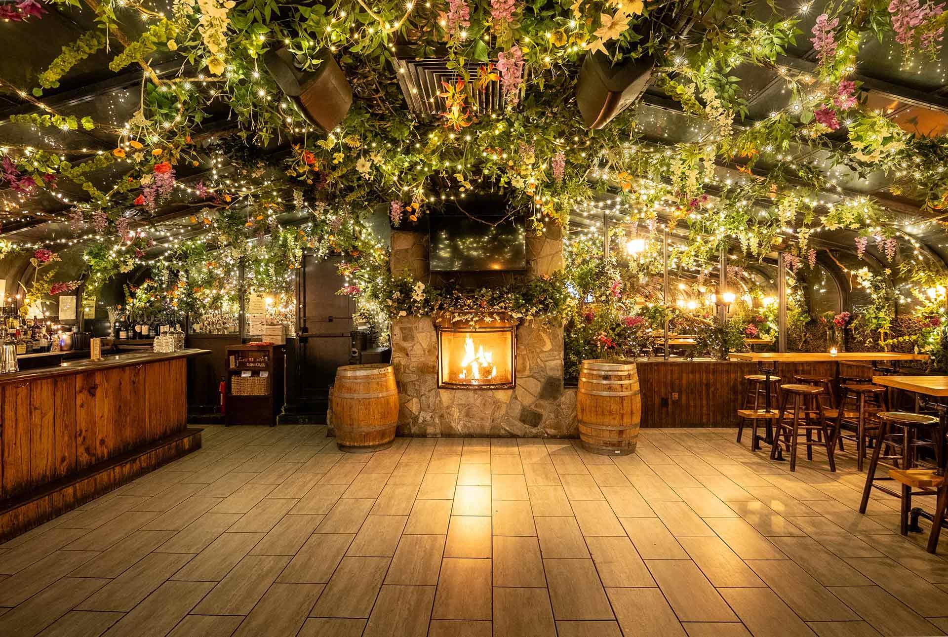A cozy bar with wooden furniture and a stone fireplace, decorated with hanging greenery, flowers, and string lights creating a warm, inviting atmosphere. Two barrels stand near the fireplace, enhancing the rustic charm.