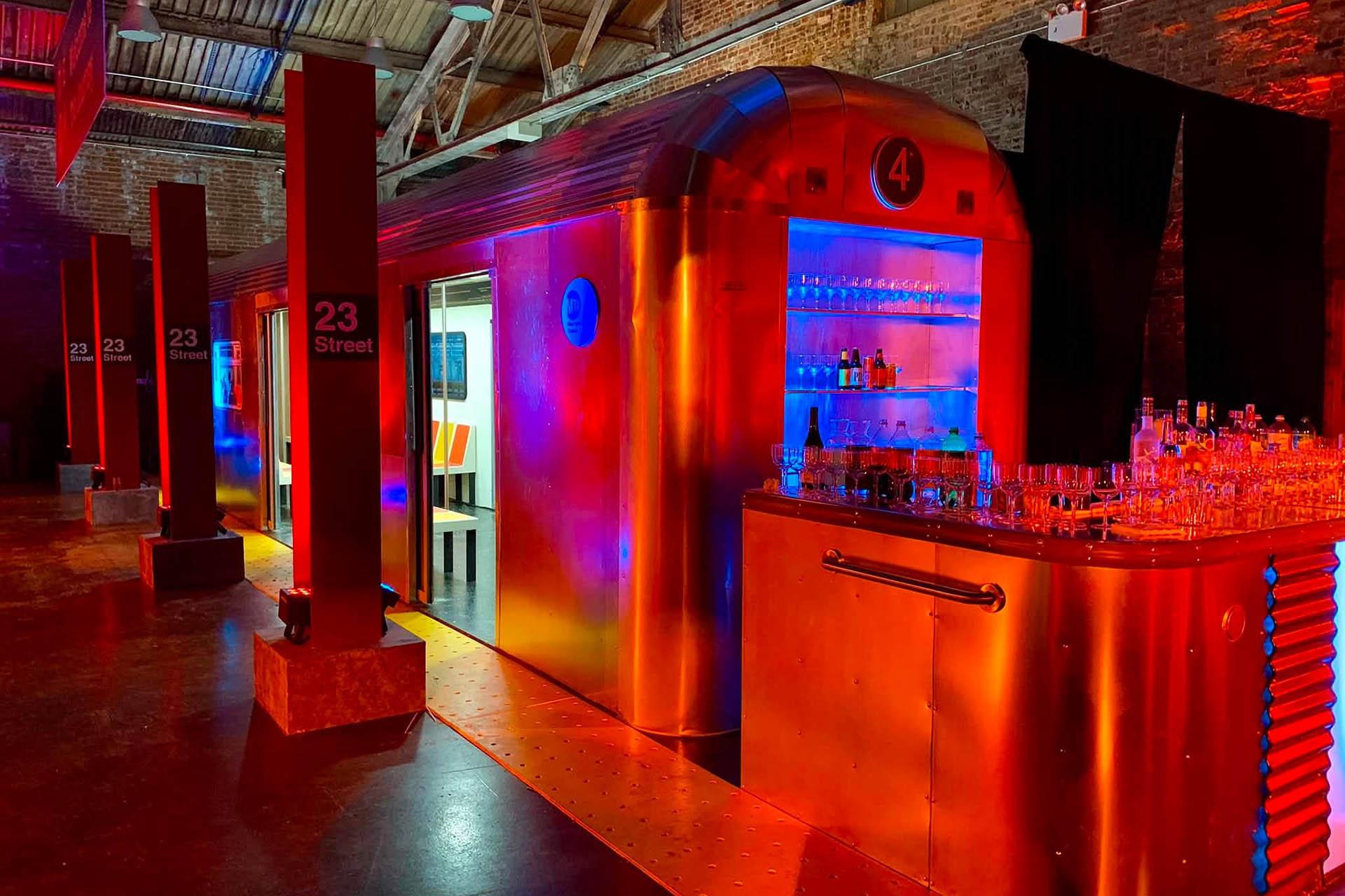 A bar setup inside a space designed to look like a vintage subway train, with neon lighting and shelves stocked with bottles and glasses, under a high industrial ceiling.