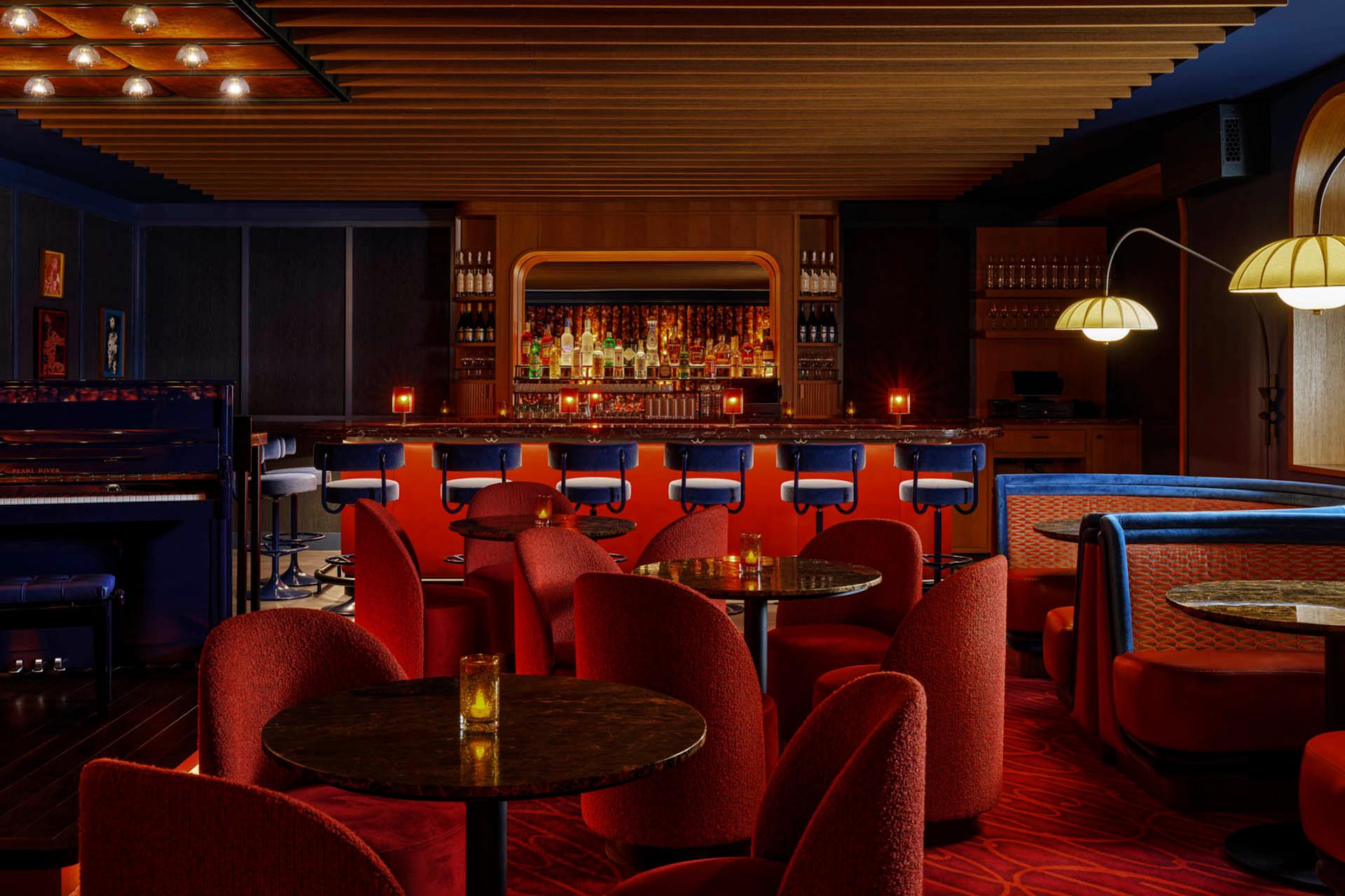 A cozy, dimly lit bar with retro red chairs and round tables. The bar is lined with blue barstools, and a variety of bottles are displayed on shelves. Warm lighting and a wooden ceiling create an inviting atmosphere. A piano is visible on the side.