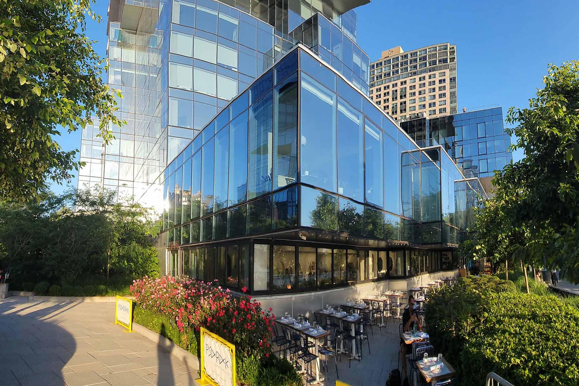 Modern glass building with reflections of the sky and surrounding structures. Patio area with outdoor seating, tables, and chairs partially visible. Lush greenery, bushes, and blooming flowers frame the scene. Bright, sunny day.