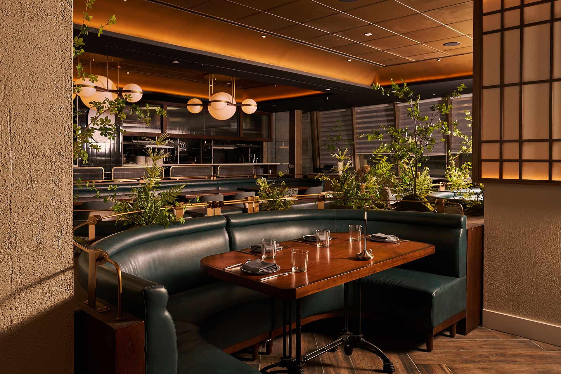 A modern restaurant interior with green leather booths, a wooden table set for four, lush green plants, and warm ambient lighting from round ceiling fixtures. The space has a cozy and inviting atmosphere.