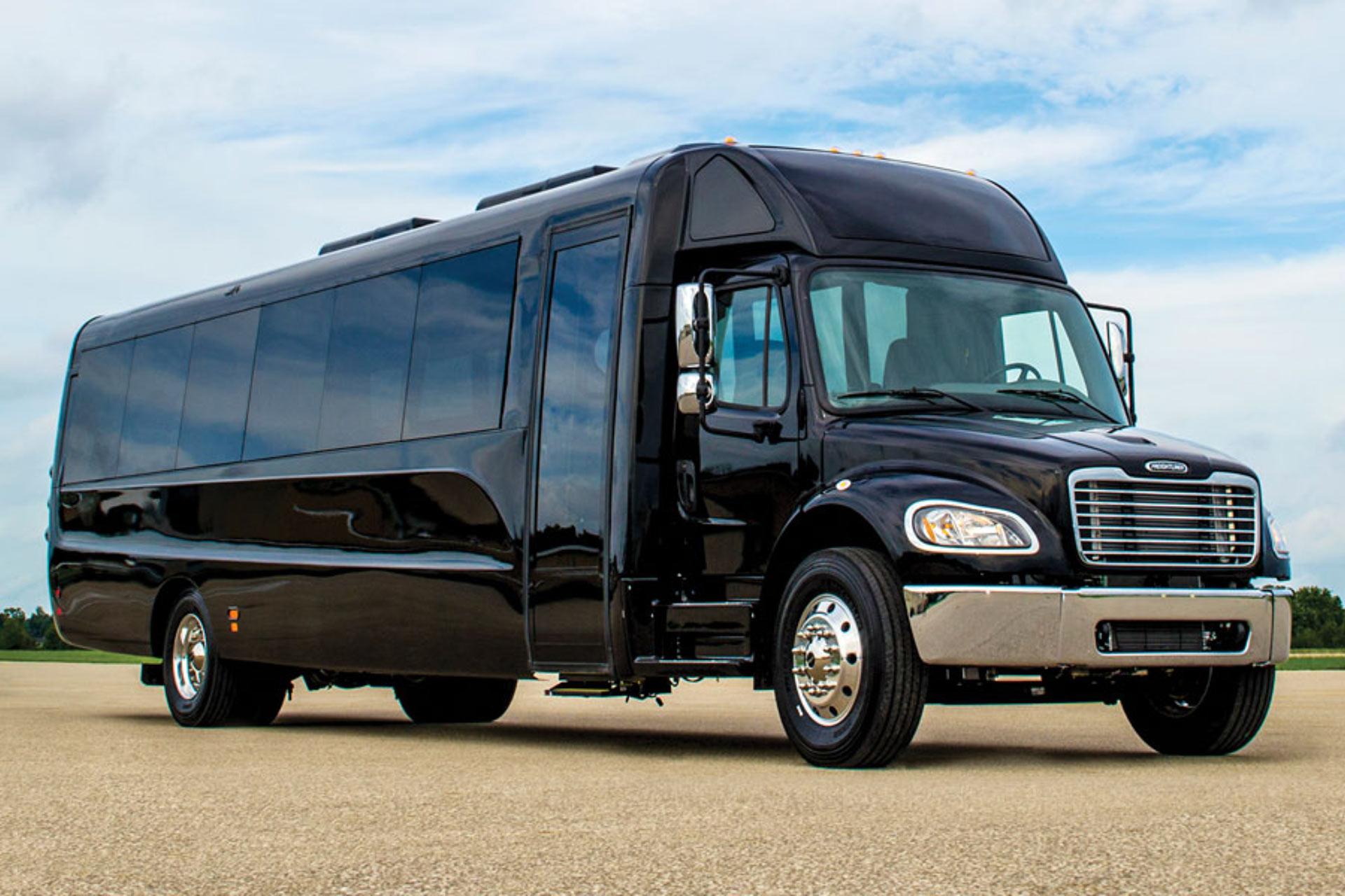 A large black luxury shuttle bus is parked on an open paved area under a partly cloudy sky, with trees and grass visible in the background.