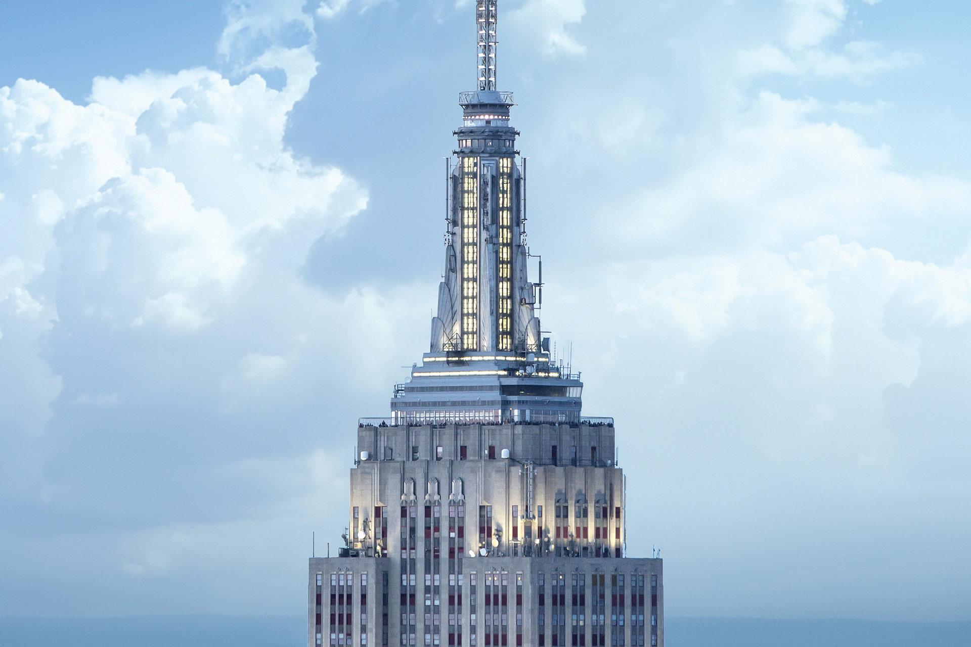 The top portion of the Empire State Building against a backdrop of bright blue sky and fluffy white clouds, showcasing its iconic spire and Art Deco architecture.