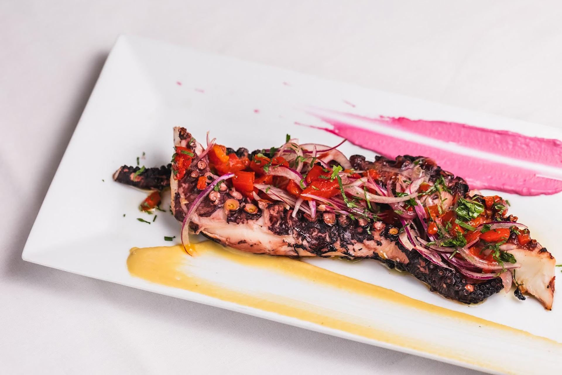 Grilled octopus tentacle topped with diced tomatoes, red onions, and herbs on a white rectangular plate, garnished with pink and yellow sauces.
