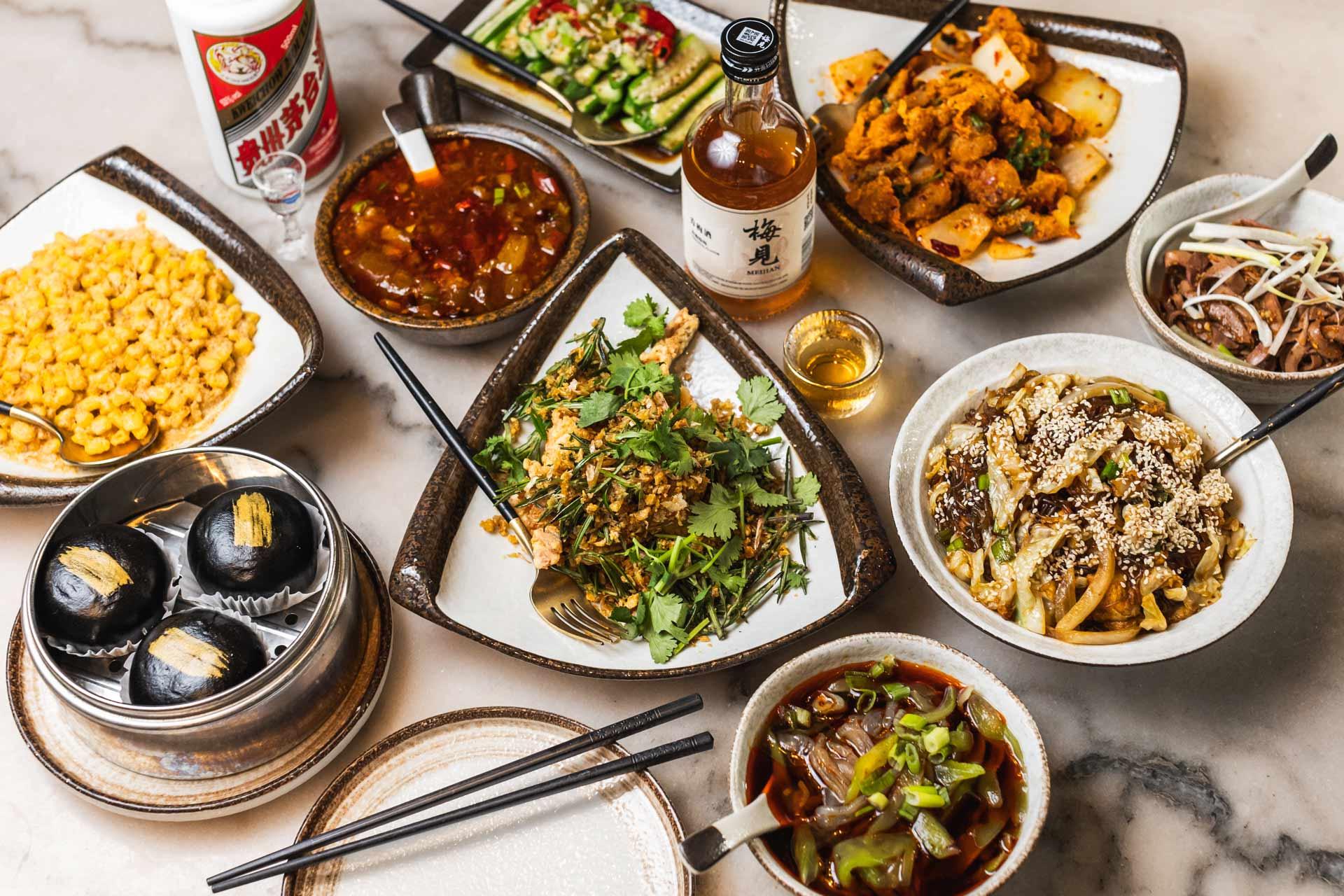 A variety of Asian dishes are artistically arranged on a table. The spread includes savory stir-fries, spicy soups, sesame-topped noodles, marinated meats, and colorful vegetable dishes, with a bottle of liquor and a glass nearby.