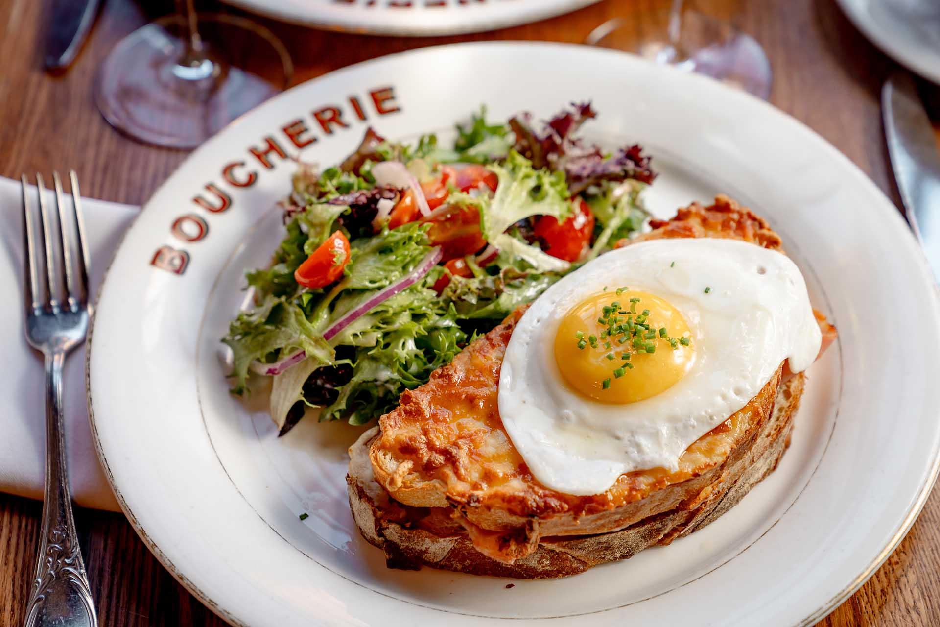 A plate with a toasted sandwich topped with melted cheese and a sunny-side-up egg, served beside a fresh mixed green salad with tomatoes and red onions.