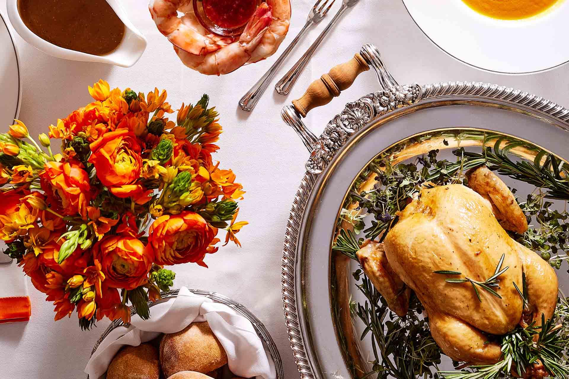 A roast chicken on a silver tray with herbs, orange flowers in a vase, bread rolls, shrimp cocktail, gravy, and soup, all arranged on a white tablecloth.