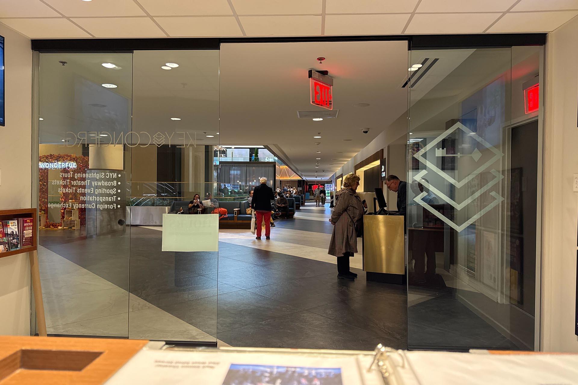 A modern building lobby seen through glass doors, with people walking and sitting. There's a reception desk on the right, ceiling lights, and an EXIT sign visible above the hallway.