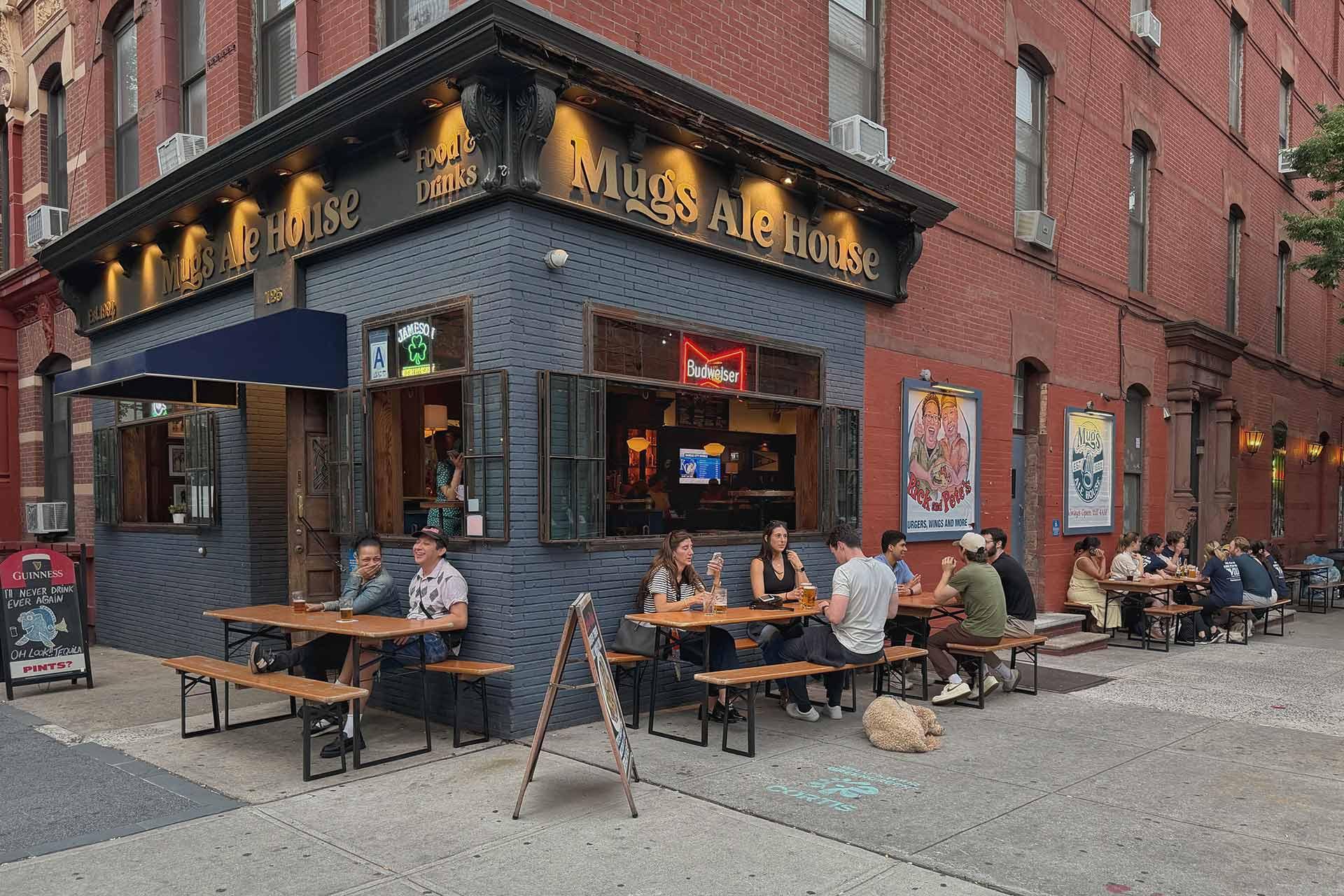 People sit at outdoor tables at Mugs Ale House, a corner bar with a dark exterior and golden signage, on a city street. Some are eating and drinking, enjoying a casual atmosphere on a cloudy day.