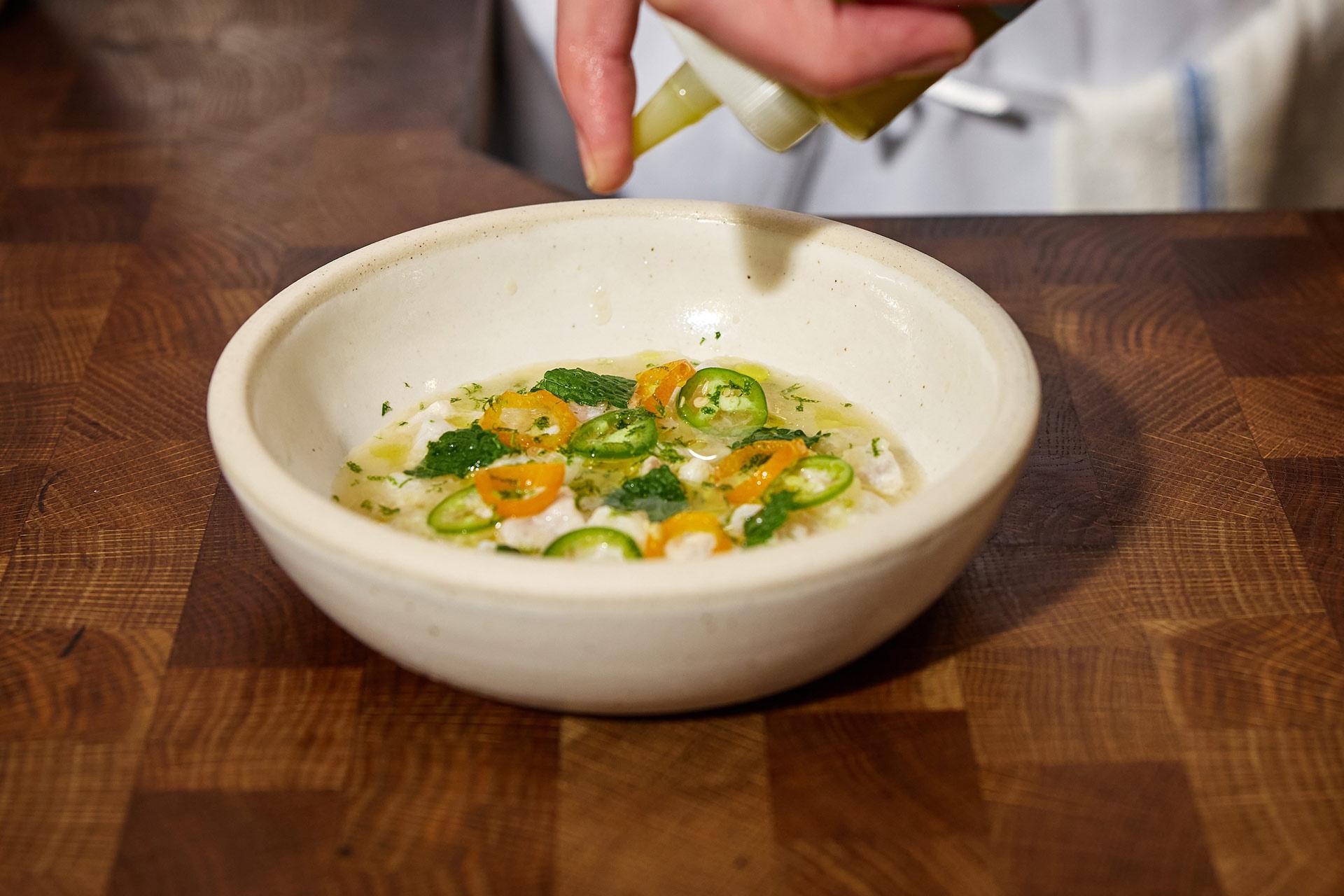 A hand drizzles oil over a bowl of ceviche garnished with sliced green and yellow chilies and fresh herbs, set on a wooden countertop.